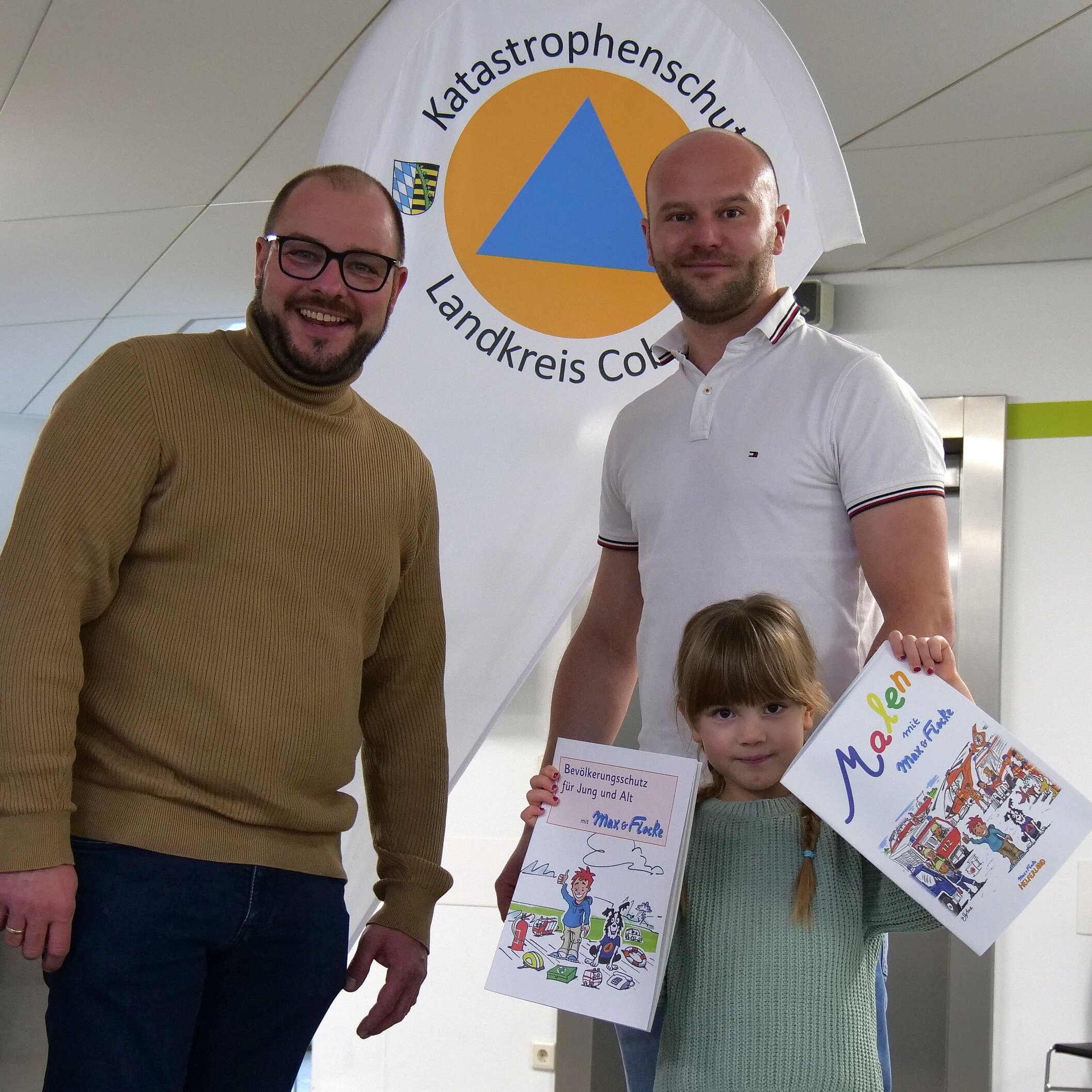 Die sechsjährige Ellie steht vorne und hält ein Mal- sowie ein Rätselbuch in die Höhe. Hinter ihr stehen Sebastian Straubel und Patrice Kolb.