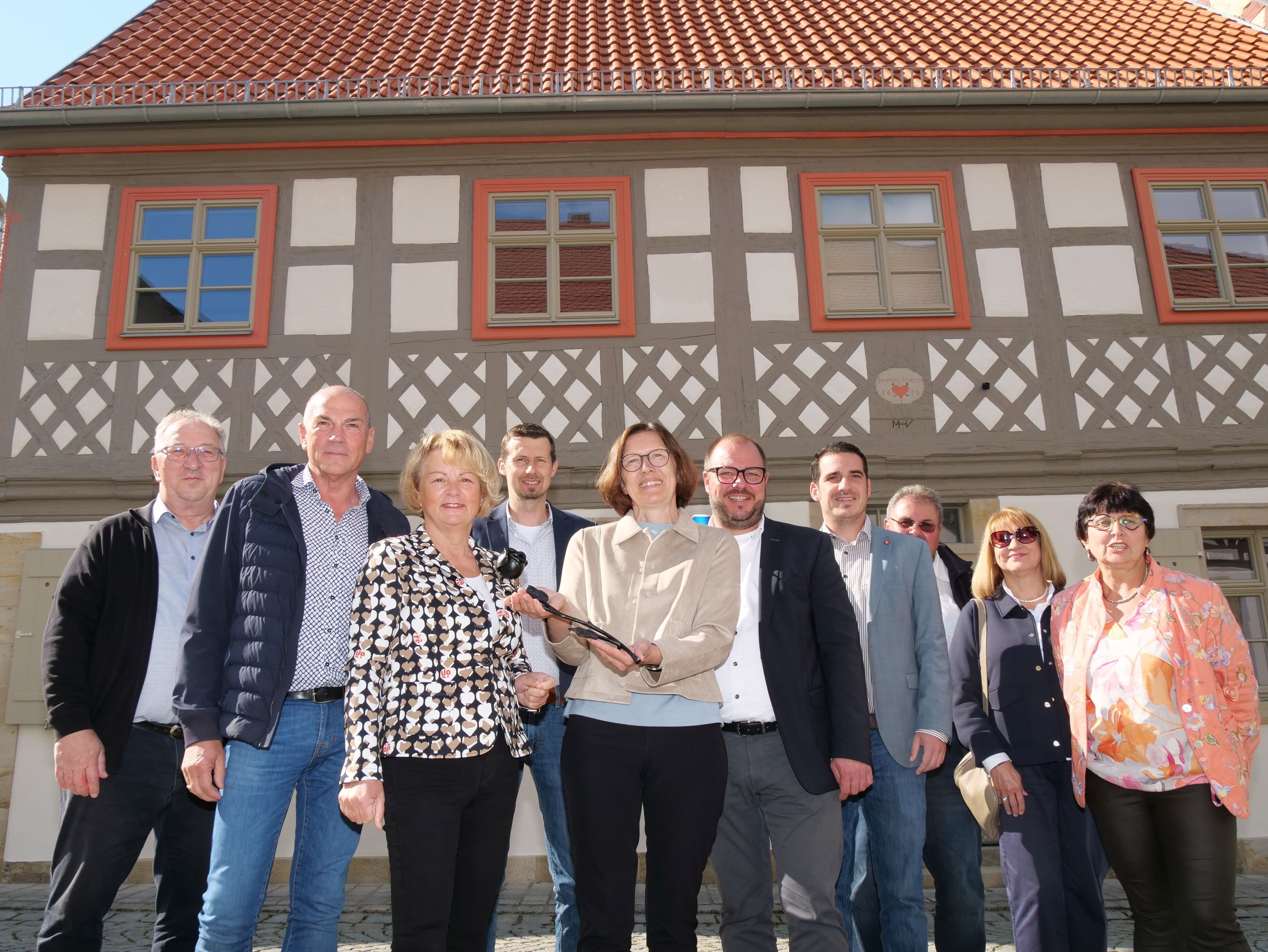 Die Familie Riedel und die Jury für die "Eiserne Rose" vor einem Fachwerkhaus in der Seßlacher Innenstadt. Das Haus ist frisch saniert, man erkennt die Holz-Sprossenfenster sowie das grau gestrichene Holz-Fachwerk.