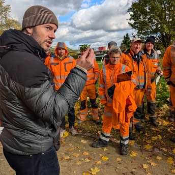 Im Vordergrund steht Referent Thomas Pickel mit einer kleinen grünen Pflanze in der Hand, ihm lauschen im Hintergrund mehrere Männer in orangefarbener Arbeitskleidung.