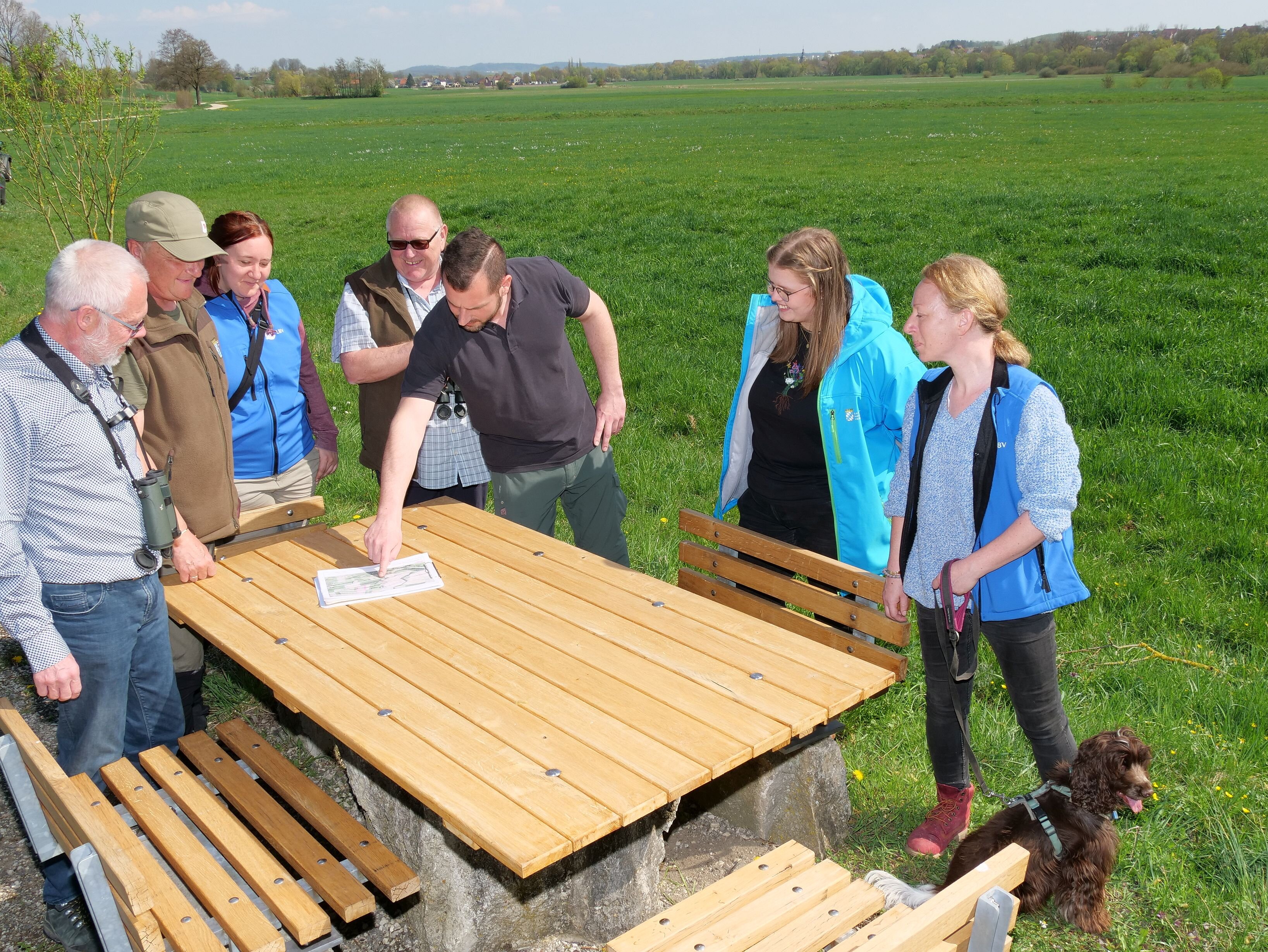 Sieben Personen stehen an einer Sitzgruppe unter freiem Himmel, auf dem Tisch liegt eine Landkarte, im Hintergrund öffnen sich die grünen Wiesen des Tales im Itzgrund.