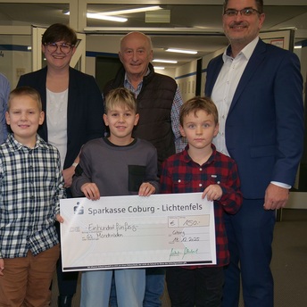 Drei Jugendliche stehen vorne und halten gemeinsam einen Spenden-Scheck, hinter ihnen stehen drei Erwachsene.