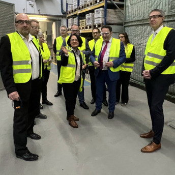 Eine Personengruppe bekleidet mit gelben Sicherheitswesten steht in einem Raum mit deckenhohen Regalen. In der Mitte steht Geschäftsführerin Brigtte Wirth, die etwas erklärt.