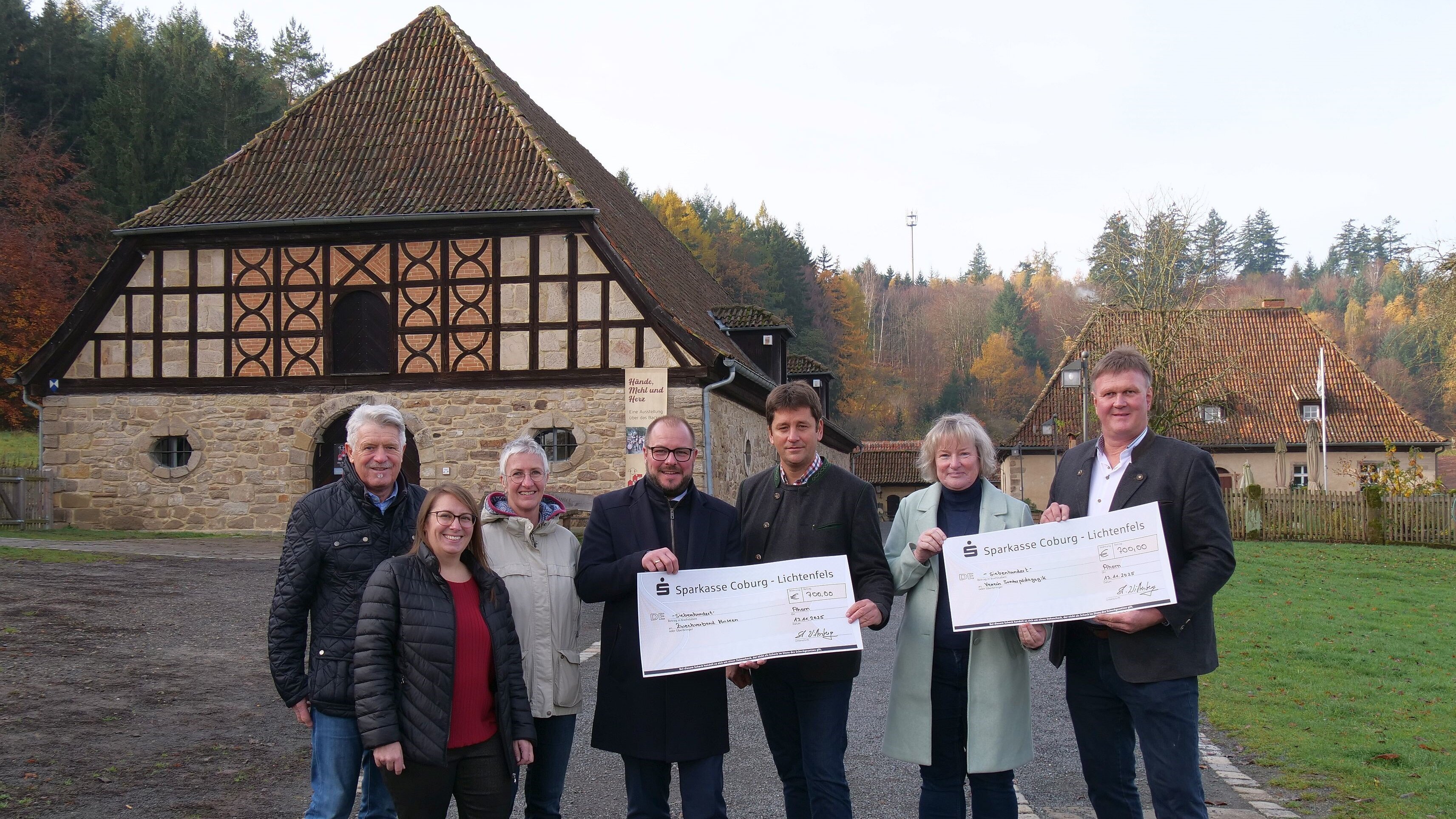Eine Gruppe mit sieben Personen steht auf dem Gelände der alten Schäferei. Die Personen halten zwei große Schecks über je 700 Euro in der Hand, im Hintergrund sieht man die alten Fachwerkgebäude der alten Schäferei.