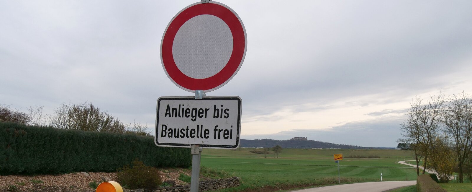 Eine Absperrbarke und eine "Durchfahrt-Verboten-Schild" sind vor einer Landstraße zu sehen. 