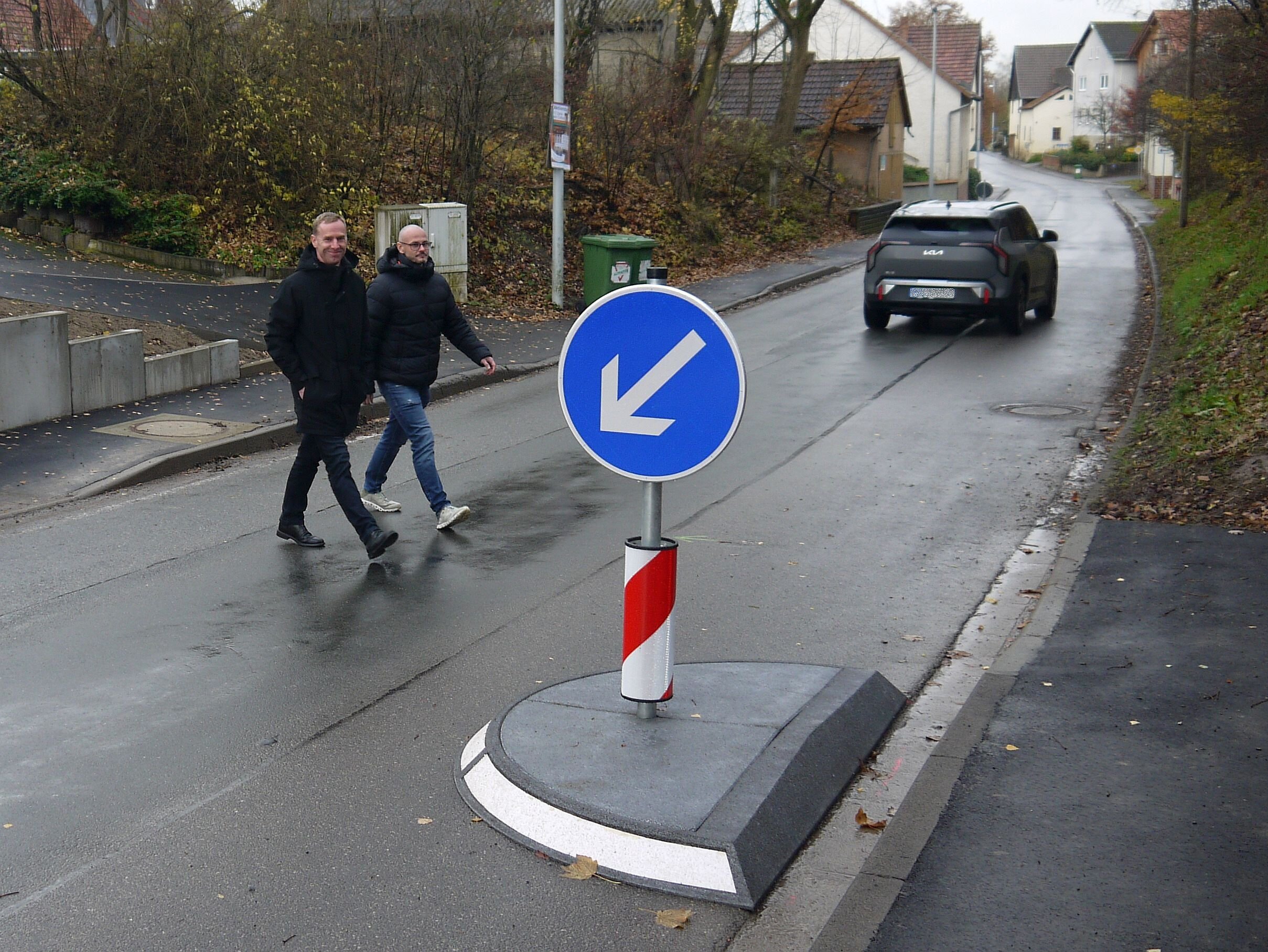 Zwei Personen überqueren eine Straße, im Hintergrund fährt ein Fahrzeug davon. Vorne sind eine Verkehrsinsel am Fahrbahnrand sowie Schild mit einem blauen Hinweispfeil zu sehen