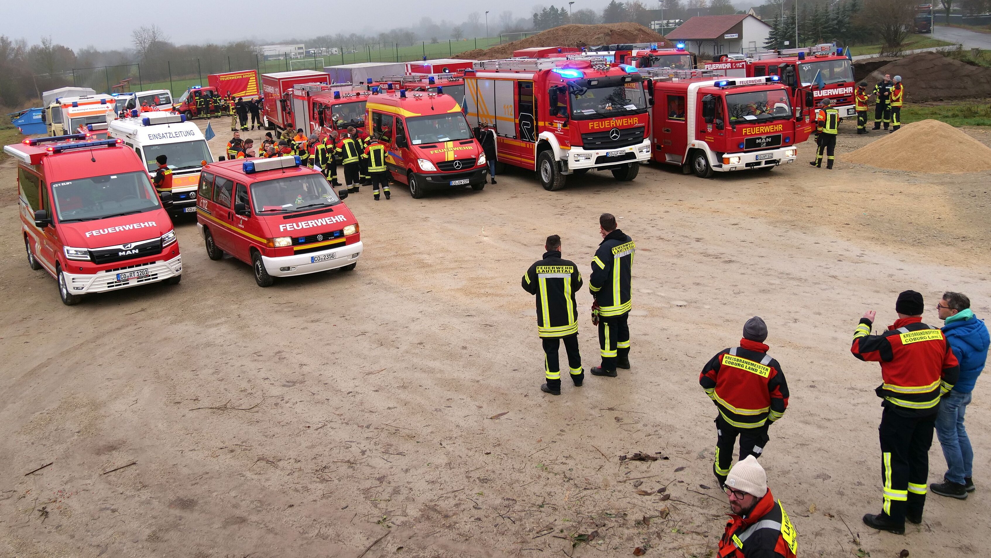 Bild von oben: Über 20 rote Einsatzfahrzeuge der Feuerwehren stehen auf einem geschotterten Parkplatz. Im Vordergrund stehen Menschen in Feuerwehr-Einsatzkleidung.