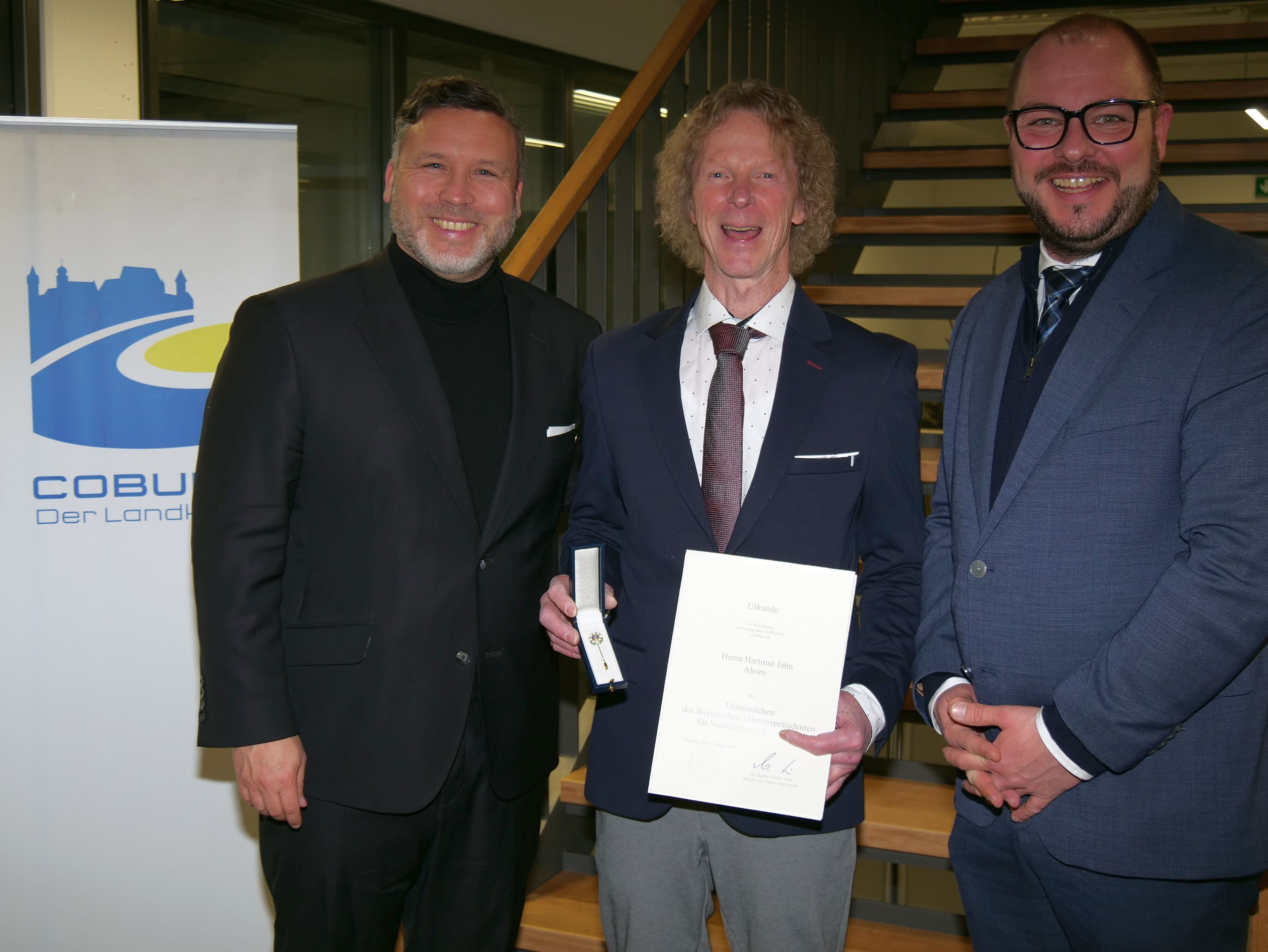 Hartmut Jahn steht lachend zwischen Sebastian Straubel und Martin Finzel. Hartmut Jahn hält dabei das Ehrenzeichen des Bayerischen Ministerpräsidenten sowie eine Urkunde in der Hand.