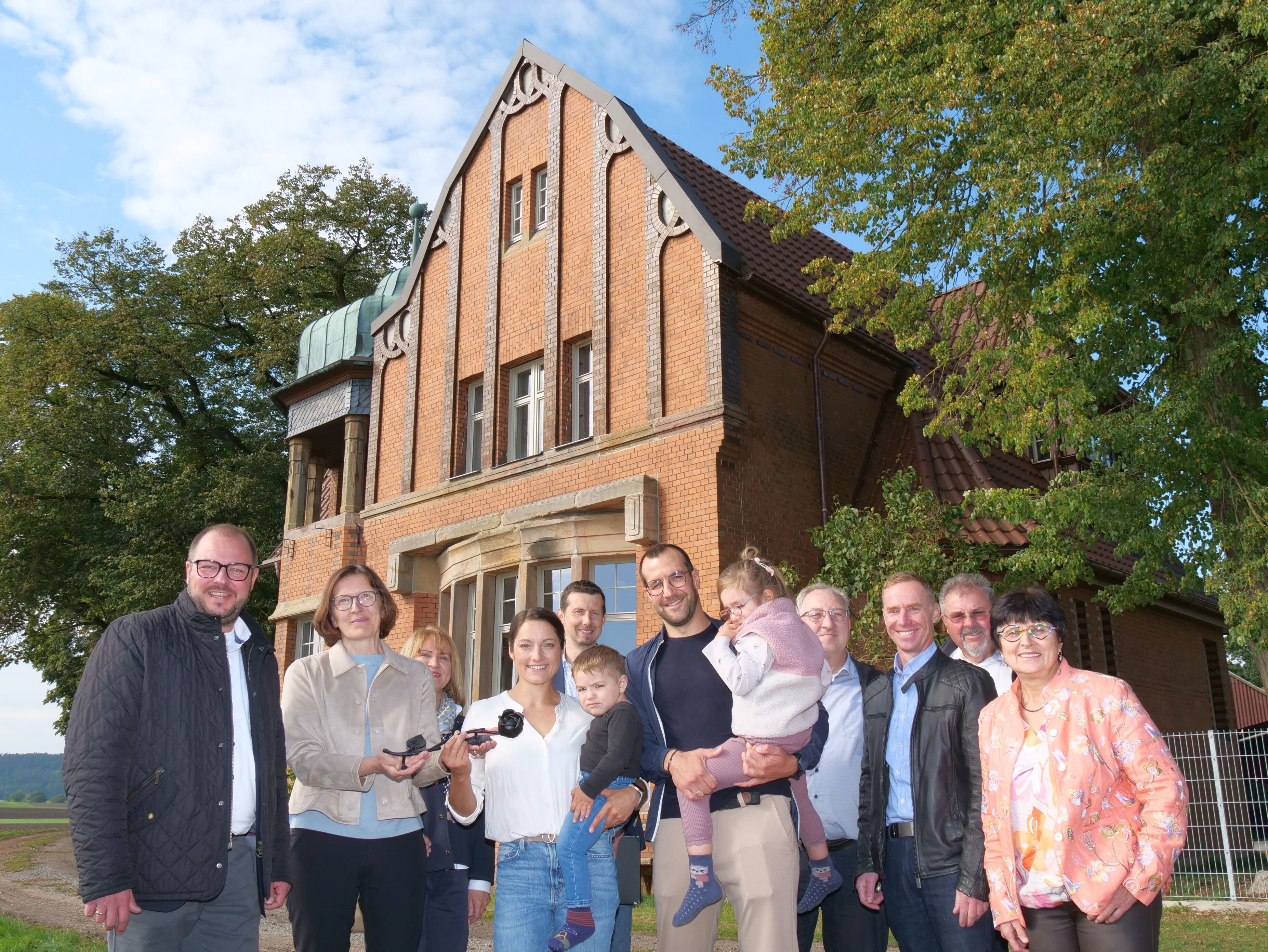 Die Jury zur Eisernen Rose und die Familie Großmannn vor dem Haus in Birkenmoor. Das ausgezeichnete Gebäude besteht aus rötlichen Backsteinen, es hat traditionelle Holz-Sprossenfenster.