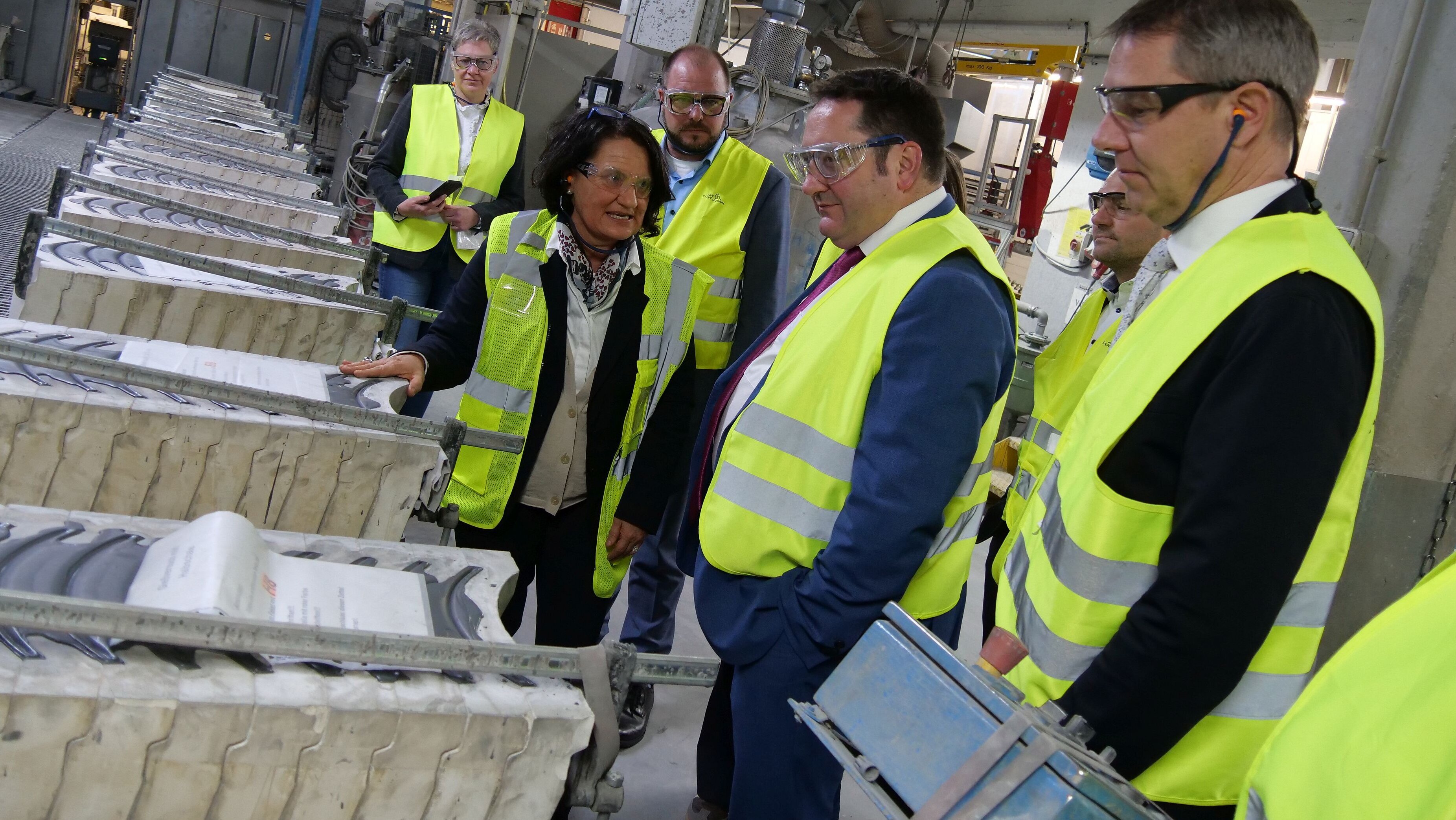 Eine Personengruppe bekleidet mit gelben Sicherheitswesten steht in einem Raum, in dem Gitterboxen mit Keramik-Platten stehen. In der Mitte steht Geschäftsführerin Brigtte Wirth, die etwas erklärt.