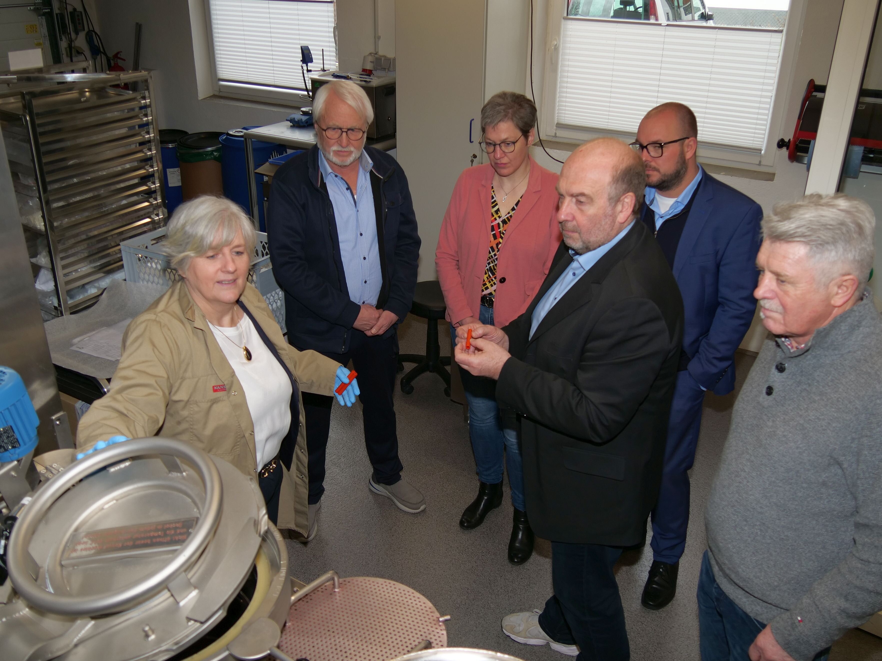 Personengruppe an einer Färbemaschine, die aussieht wie eine große Waschmaschine. Eine Frau steht links und öffnet eine Klappe des Färbebehälters.