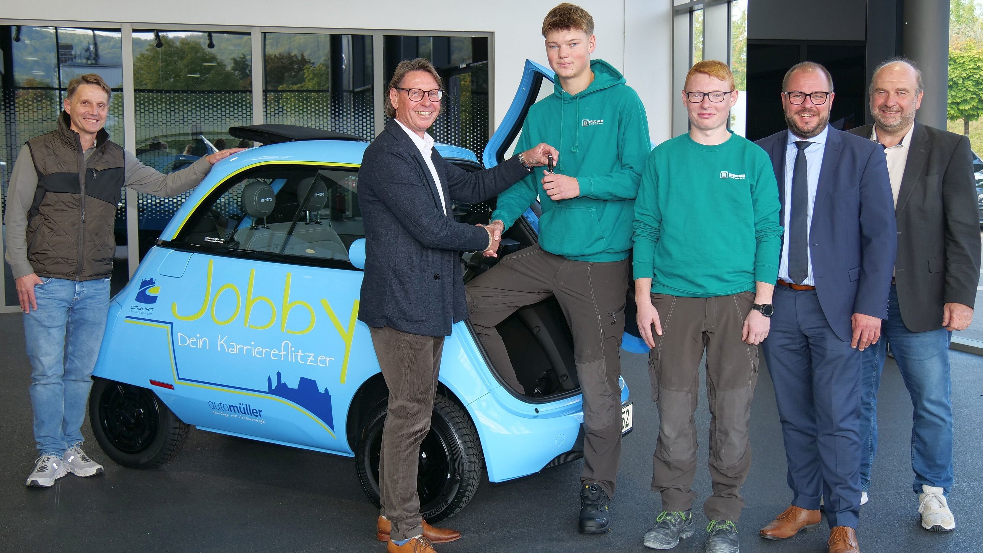 Udo Brückner, Thilo Bohl, Adrian Latyschev, Jakob Schmittlutz, Sebastian Straubel und Martin Schmitz steht vor dem himmelblau lackierten Jobby-Fahrzeug. Thilo Bohl überreicht dabei an Adrian Latyschev die Autoschlüssel für das Fahrzeug.