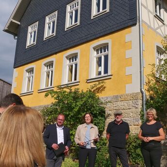 Die Jury des Denkmalschutz-Preises "Eiserne Rose" steht vor einem alten Bauernhaus, die Fassade ist gelb gestrichen, die oberen Stockwerke sind mit schwarzem Schiefer verkleidet.