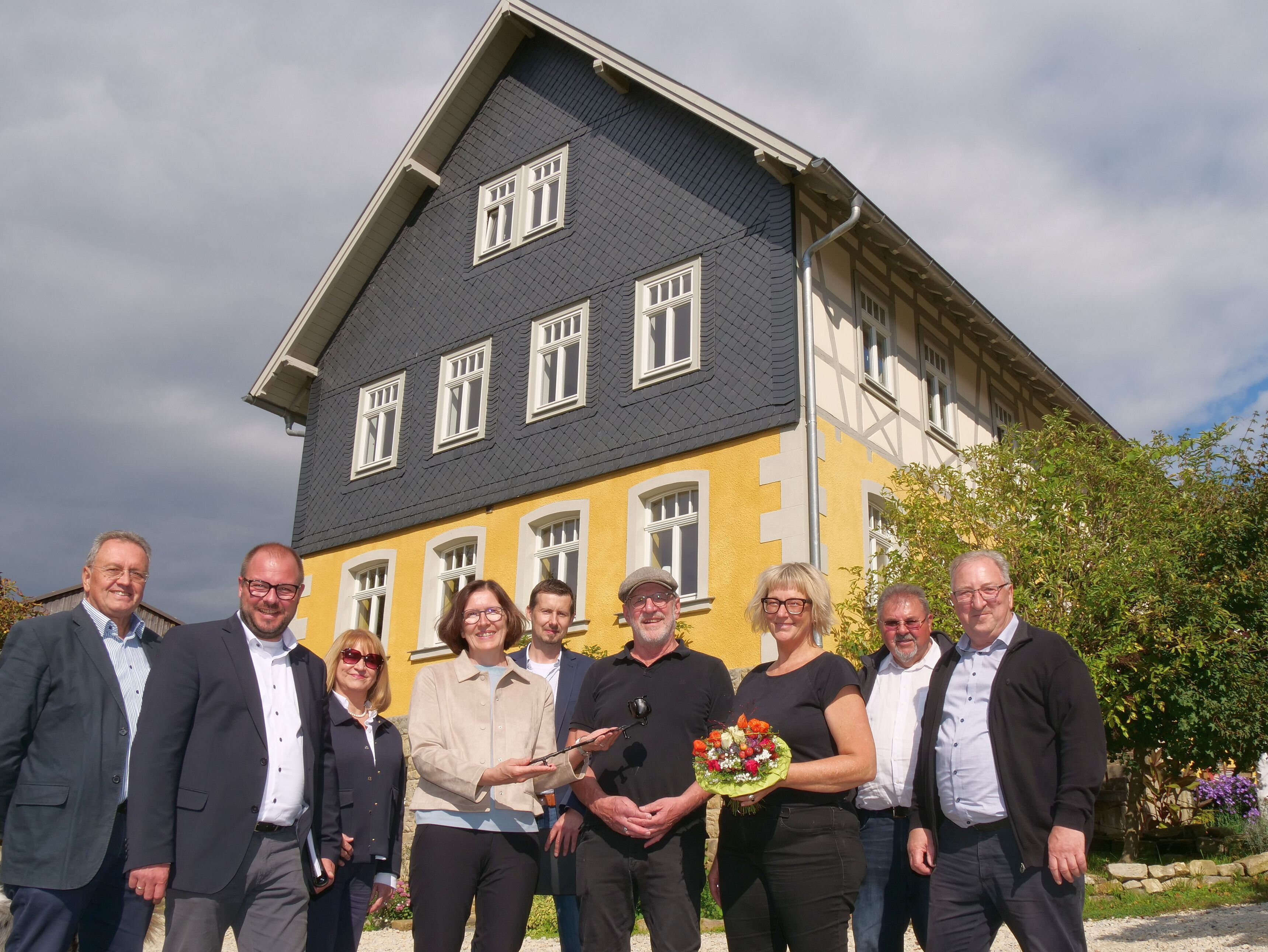Die Familie Benzel-Weyh und die Jury für die "Eiserne Rose" stehen vor dem prämierten Bauernhaus. Das frisch sanierte Haus ist gelb gestrichen, hat weiße Sprossenfenster und die oberen beiden Stochwerke sind mit grauem Schiefer verkleidet.