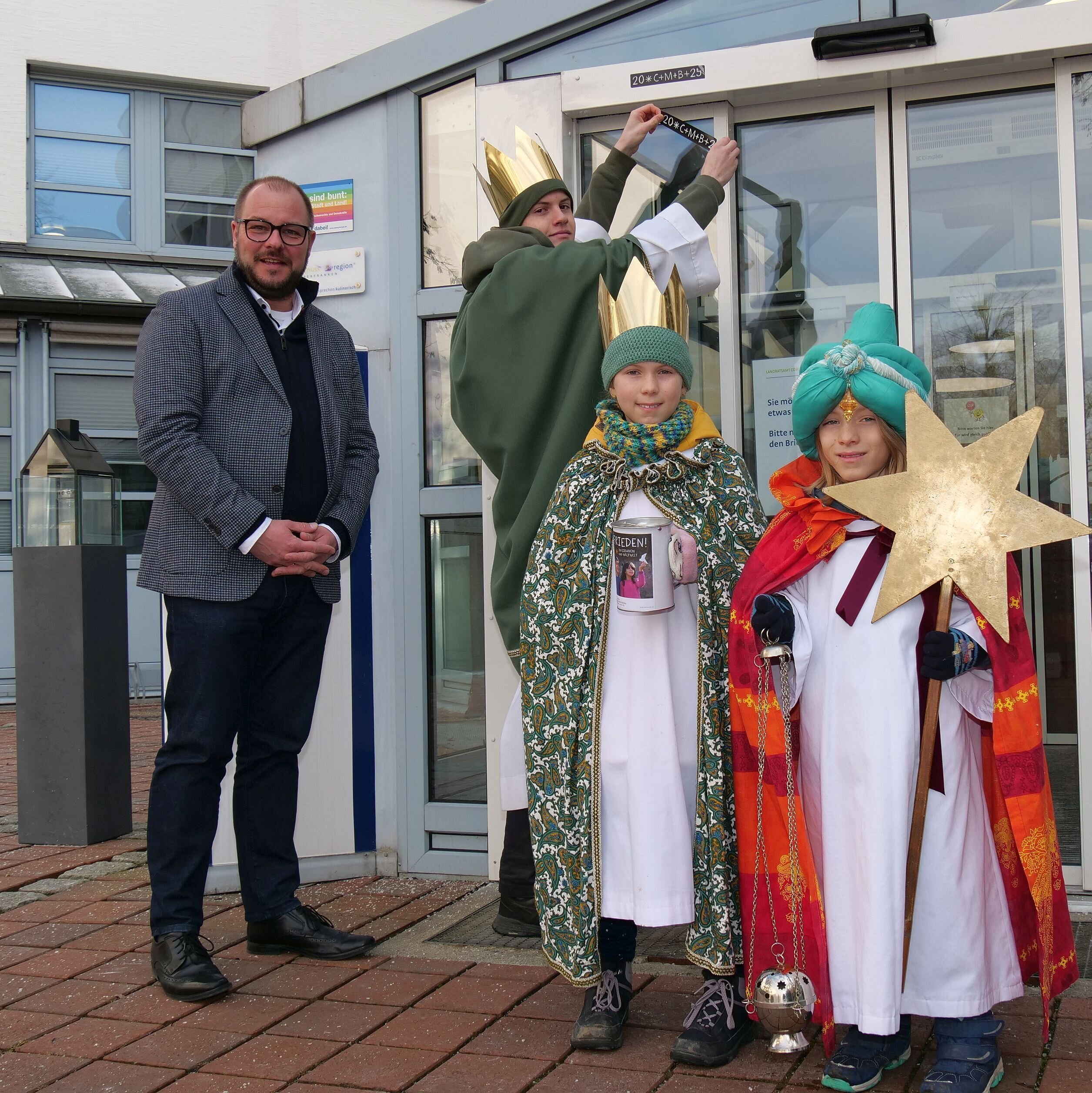 Drei Sternsinger und Landrat Sebastian Straubel stehen vor dem Haupteingang am Landratsamt Coburg. Einer der Sternsinger befestigt den Aufkleber mit dem Segensspruch über der Tür.