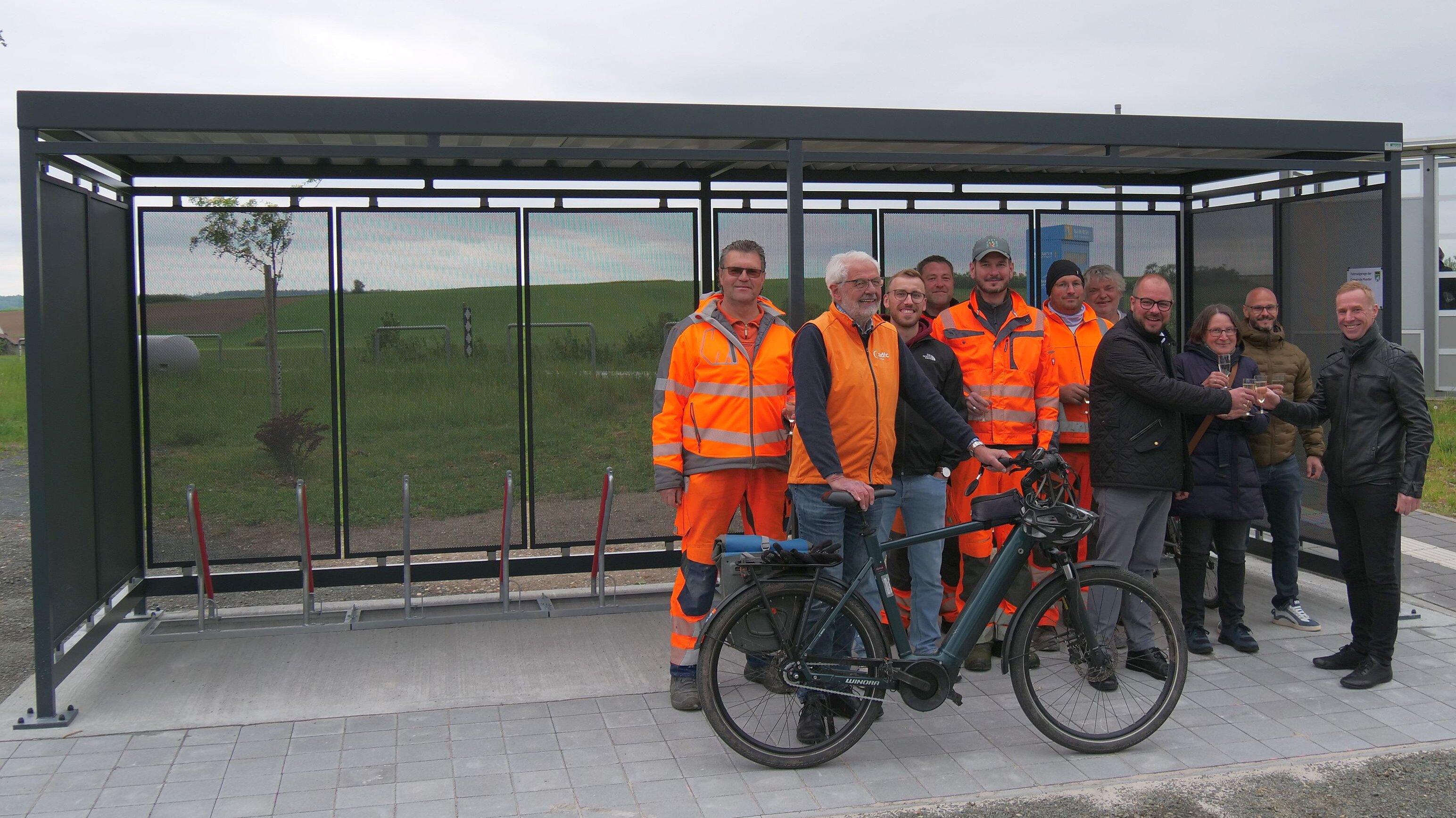  Eine größere Personengruppe, darunter Männer mit orangefarbener Arbeitskleidung, stehen vor einer überdachten Abstellanlage für Fahrräder. Einer der Männer hat ein Fahrrad dabei.