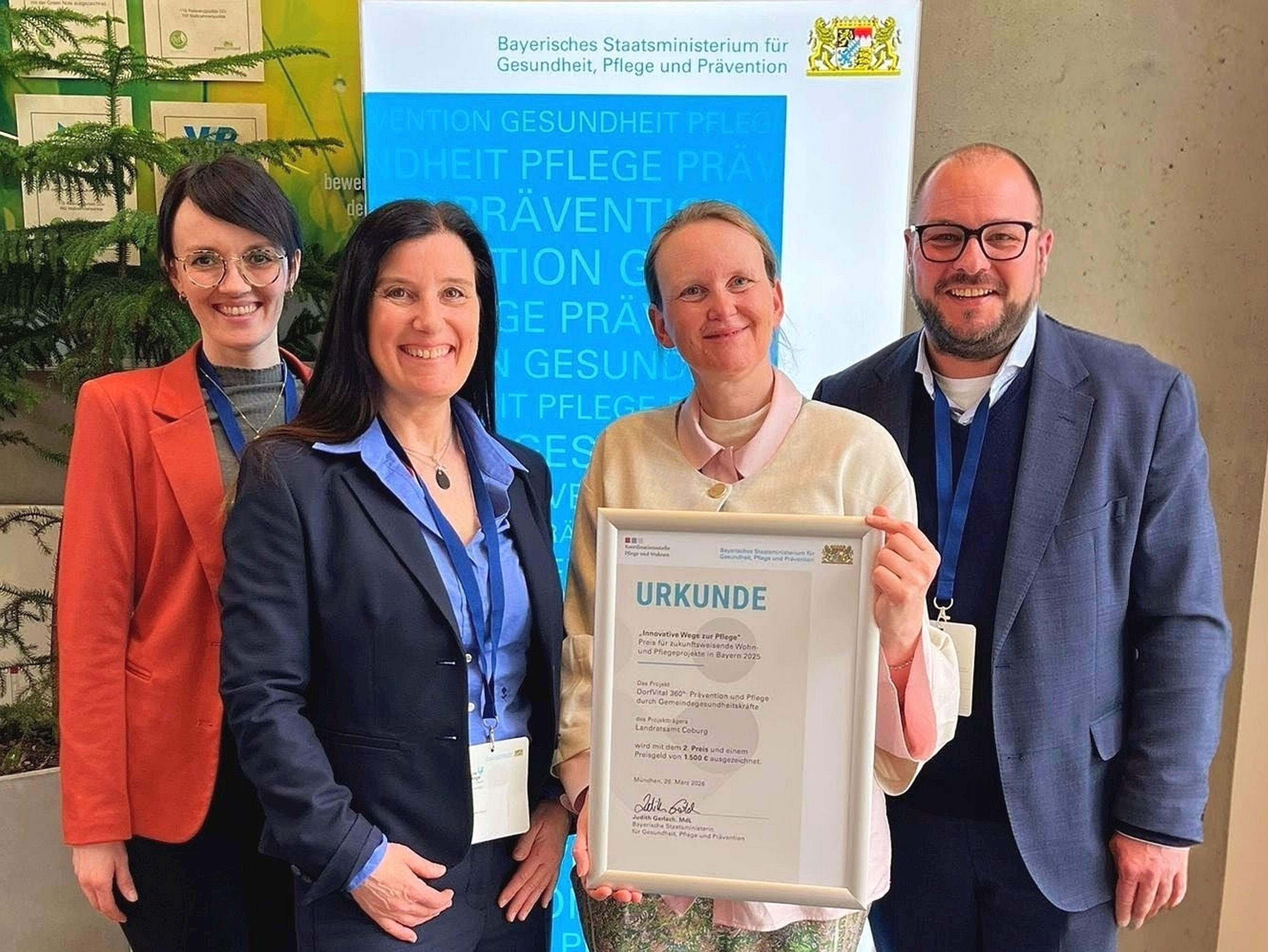 anessa Kaiser, Anja Zietz, Kathrin Jouma und Sebastian Straubel stehen vor einem Banner des Freistaats Bayern mit der Urkunde des Wettbewerbes "Innovative Pflege" in der Hand.