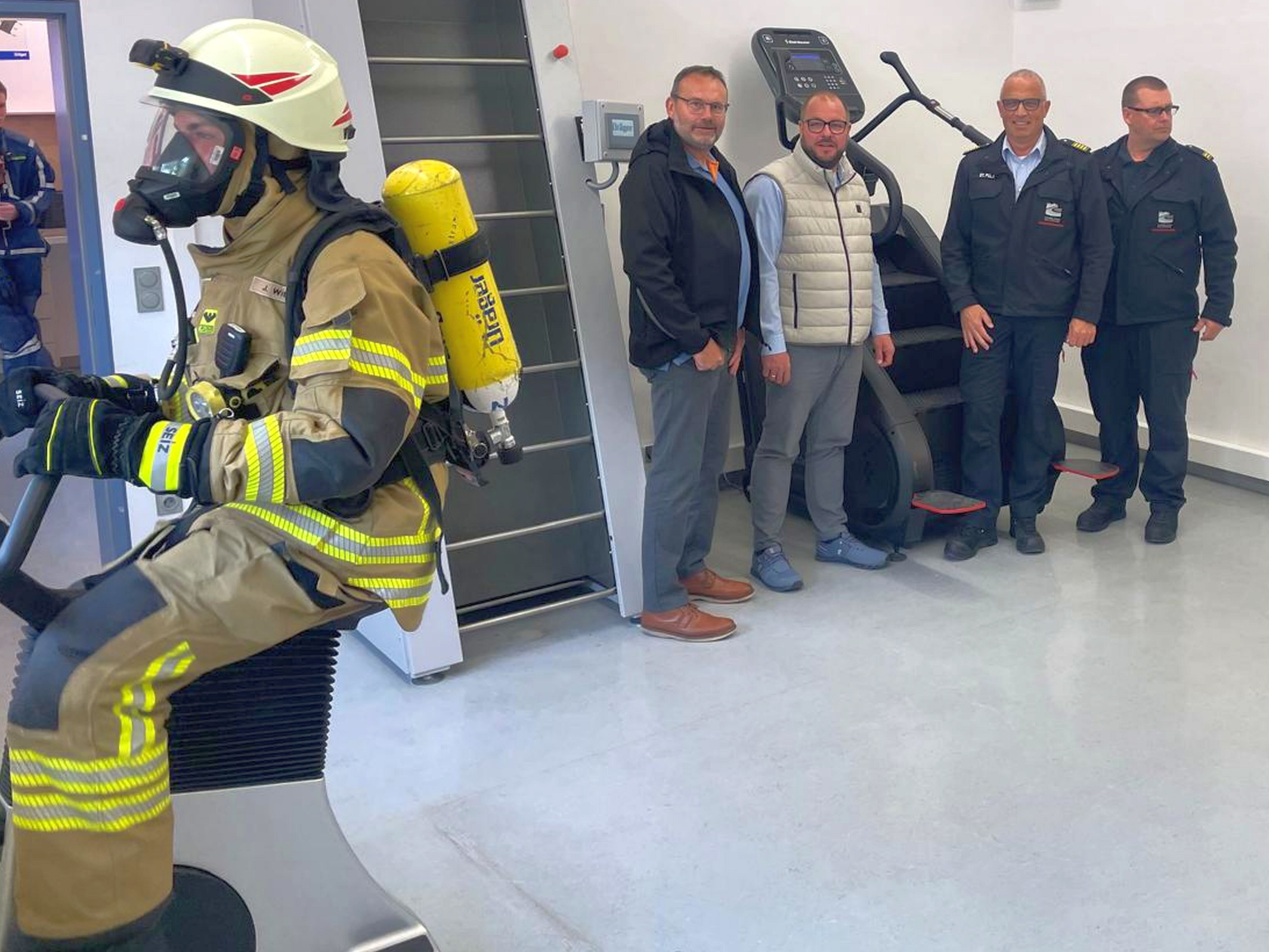 Ein Feuerwehrmann mit Atemschutzausrüstung trainiert auf einem Fahrradergometer. Mehrere Personen in Zivil- und Feuerwehr-Kleidung schauen ihm zu.