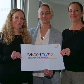 Drei Frauen stehen in einem Büro und halten gemeinsam ein Schild, auf dem Mohritz steht.