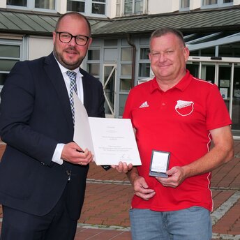 Landrat Sebastian Straubel steht links und überreicht vor dem Landratsamt Coburg an Wolfgang Rößner eine Urkunde.