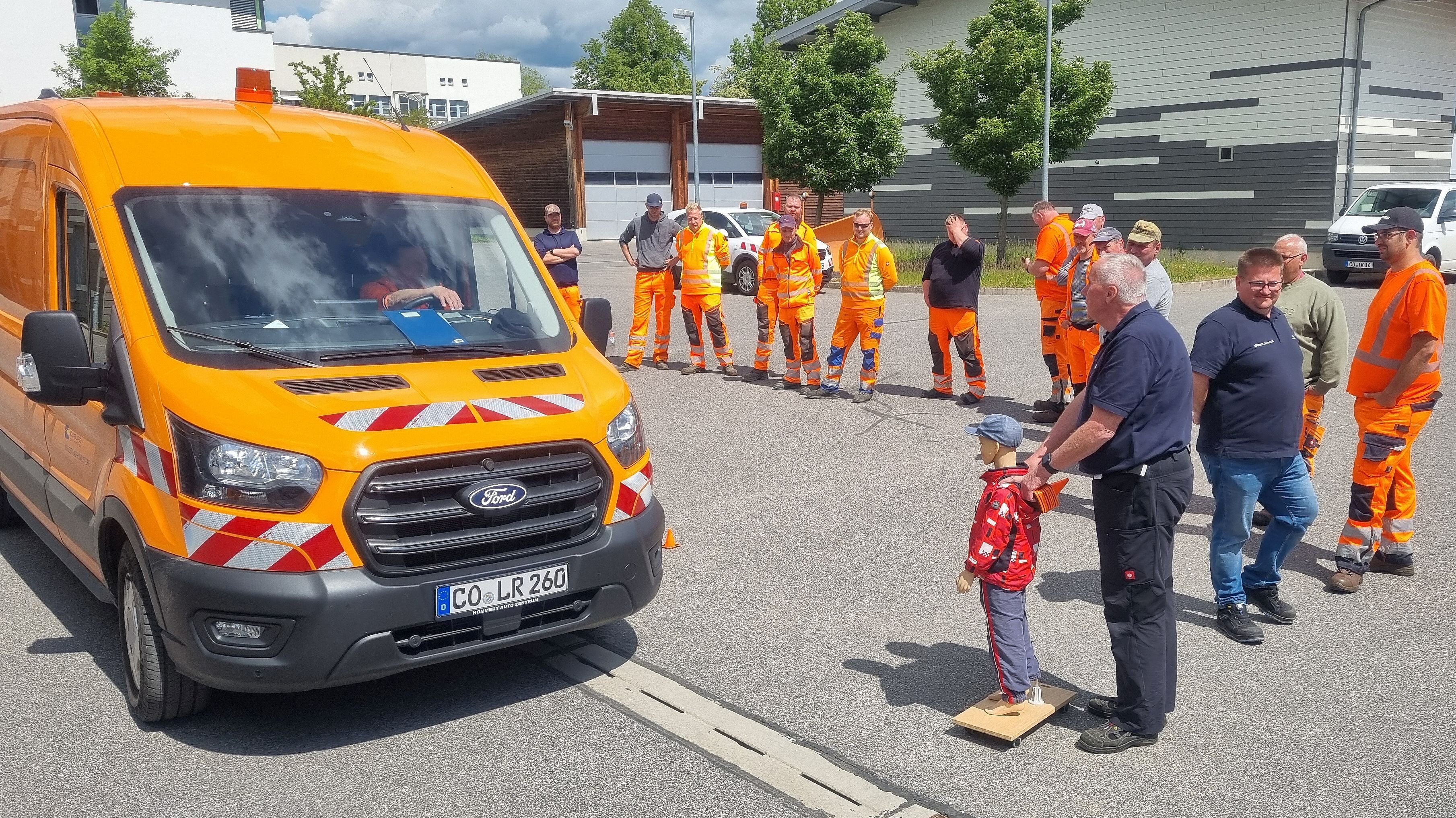 Zwei Männer in blauen Shirt schieben eine Kinderpuppe in den toten Winkel vor einem orangefarbenen Kleintransporter. Eine größere Gruppe von Männern in orangefarbener Arbeitskleidung verfolgt das Geschehen aufmerksam.