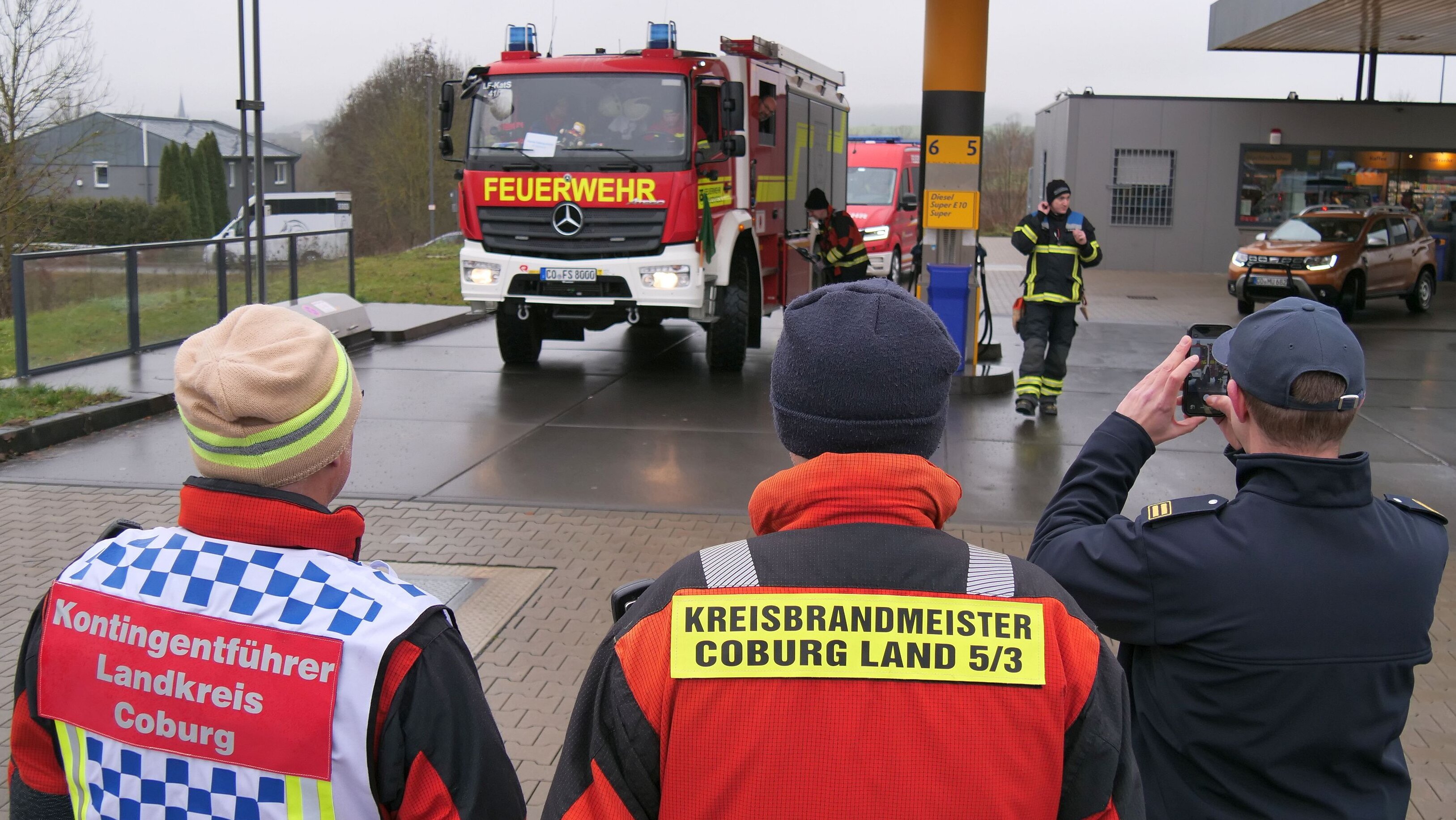 Drei Personen mit Feuerwehr-Einsatzkleidung schauen zu, wie ein Feuerwehr-Fahrzeug an einer Tankstelle Kraftstoff tankt.