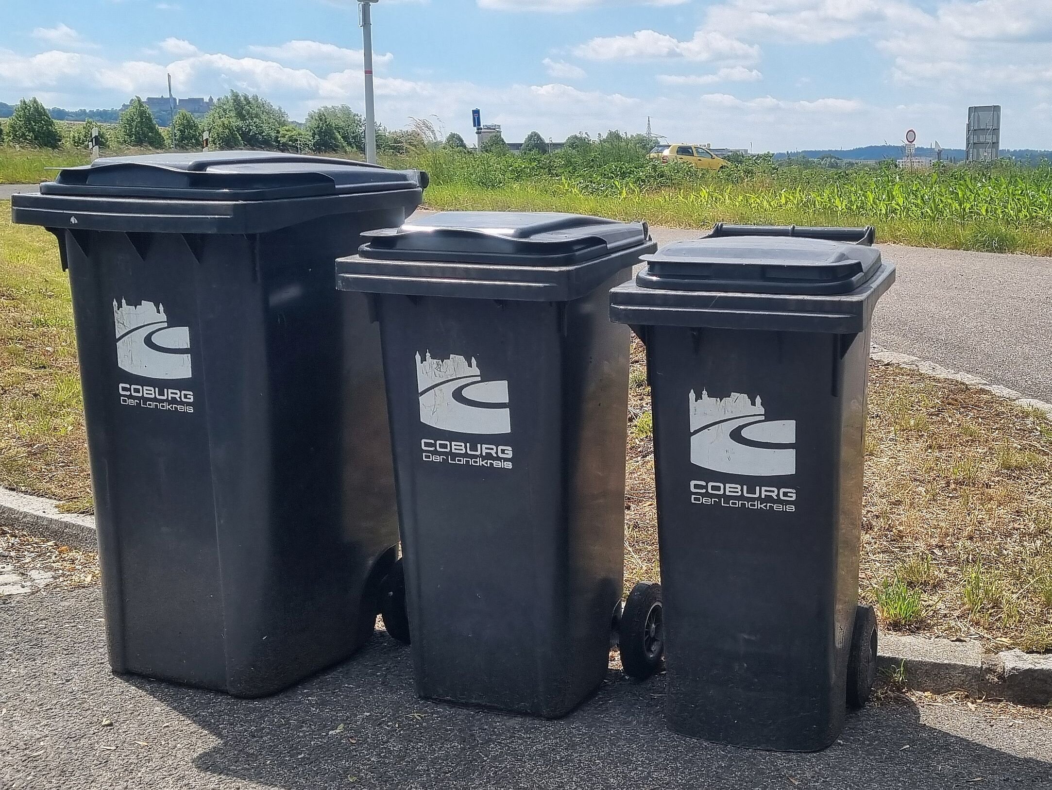 Drei Mülltonnen mit dem Logo des Landkreises Coburg stehen am Straßenrand. Im Hintergrund ist am Horizont die Veste Coburg zu sehen.