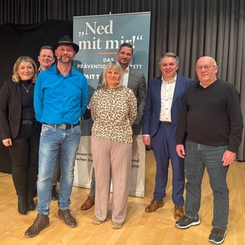 Eine Gruppe mit sieben Personen steht vor einem Roll-Up, auf dem steht "Ned mit mir"