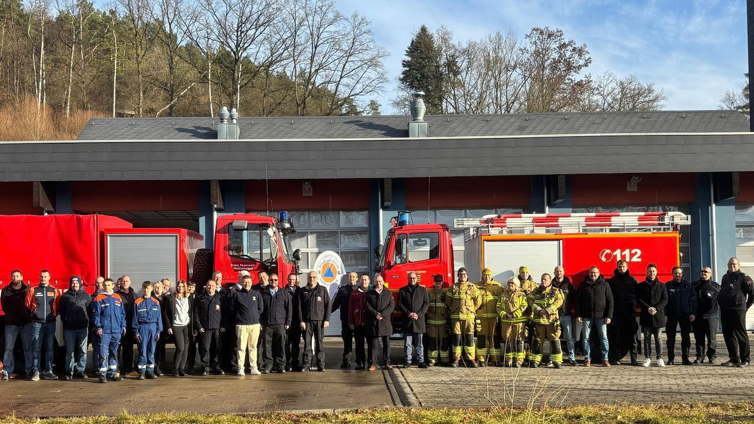 Gut zwei Dutzend Feuerwehr-Aktive in Einsatzkleidung stehen zum Gruppenbild versammelt vor zwei roten Feuerwehrfahrzeugen.