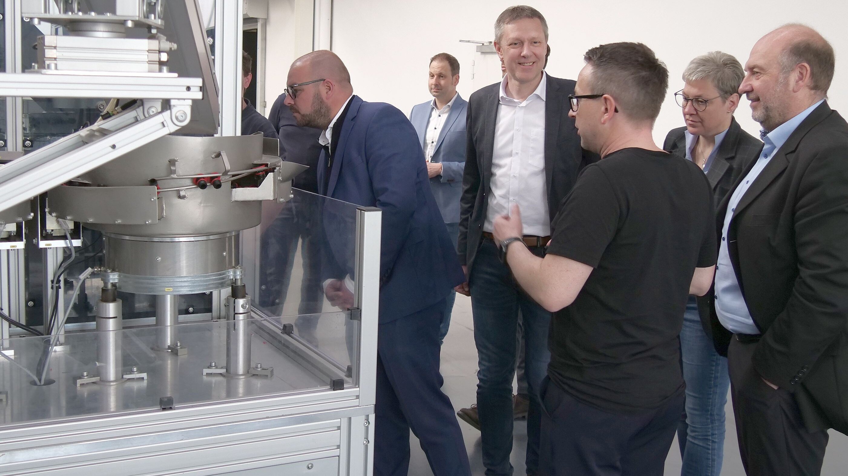 Eine Personengruppe steht vor einer Spritzguss-Maschine. Link steht Landrat Sebastian Straubel, der ins Innere der Maschine schaut.
