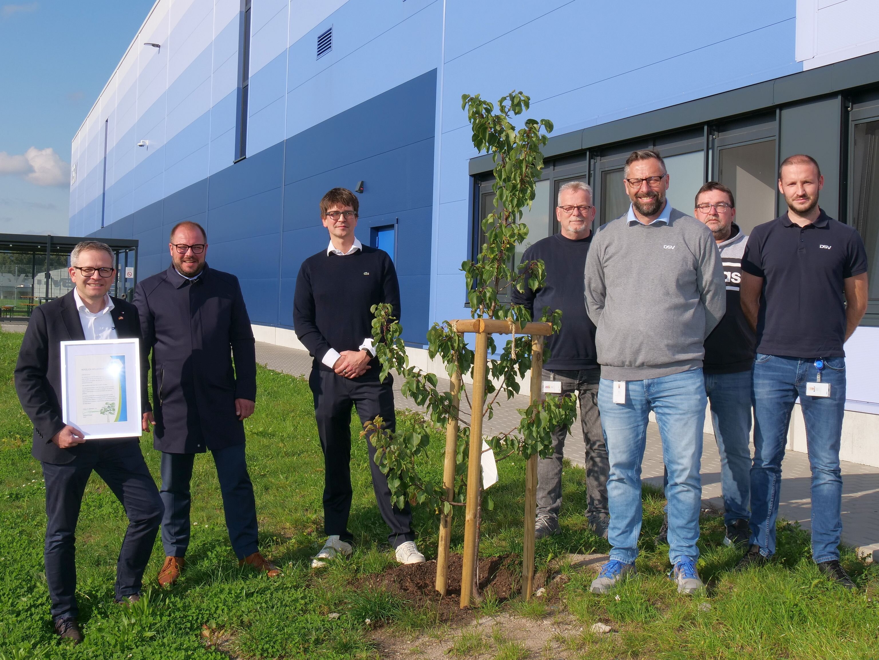 Sieben Personen stehen vor dem Firmengebäude der DSV-Spedition neben einem frisch gepflanzten Baum. Ganz links steht ein Mann mit einer Urkunde in Händen.