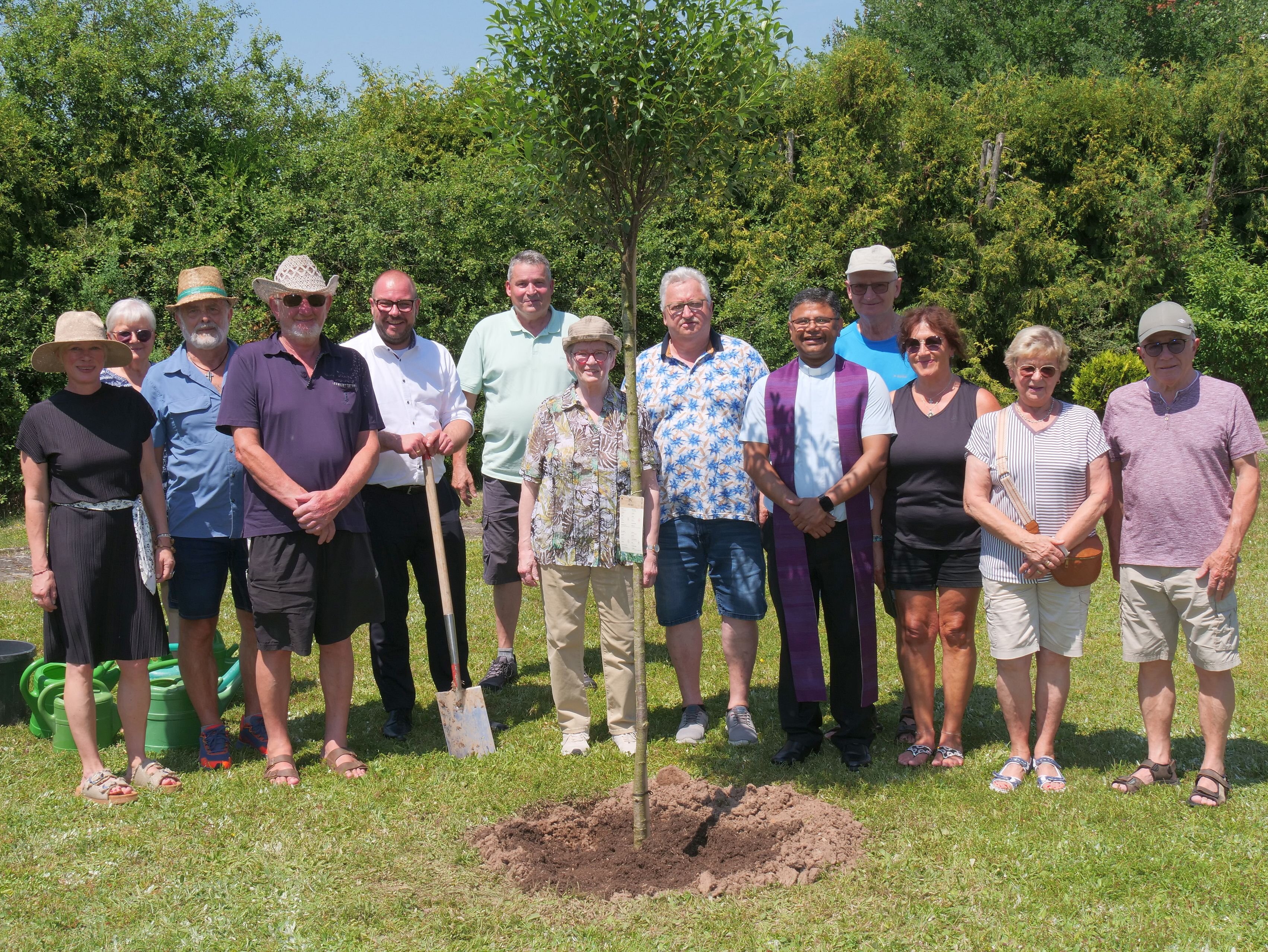 Ein Gruppenfoto mit gut einem Dutzend Personen, die - teils mit Gartenwerkzeug ausgestattet - vor einem gut drei Meter hohen jungen Baum stehen.