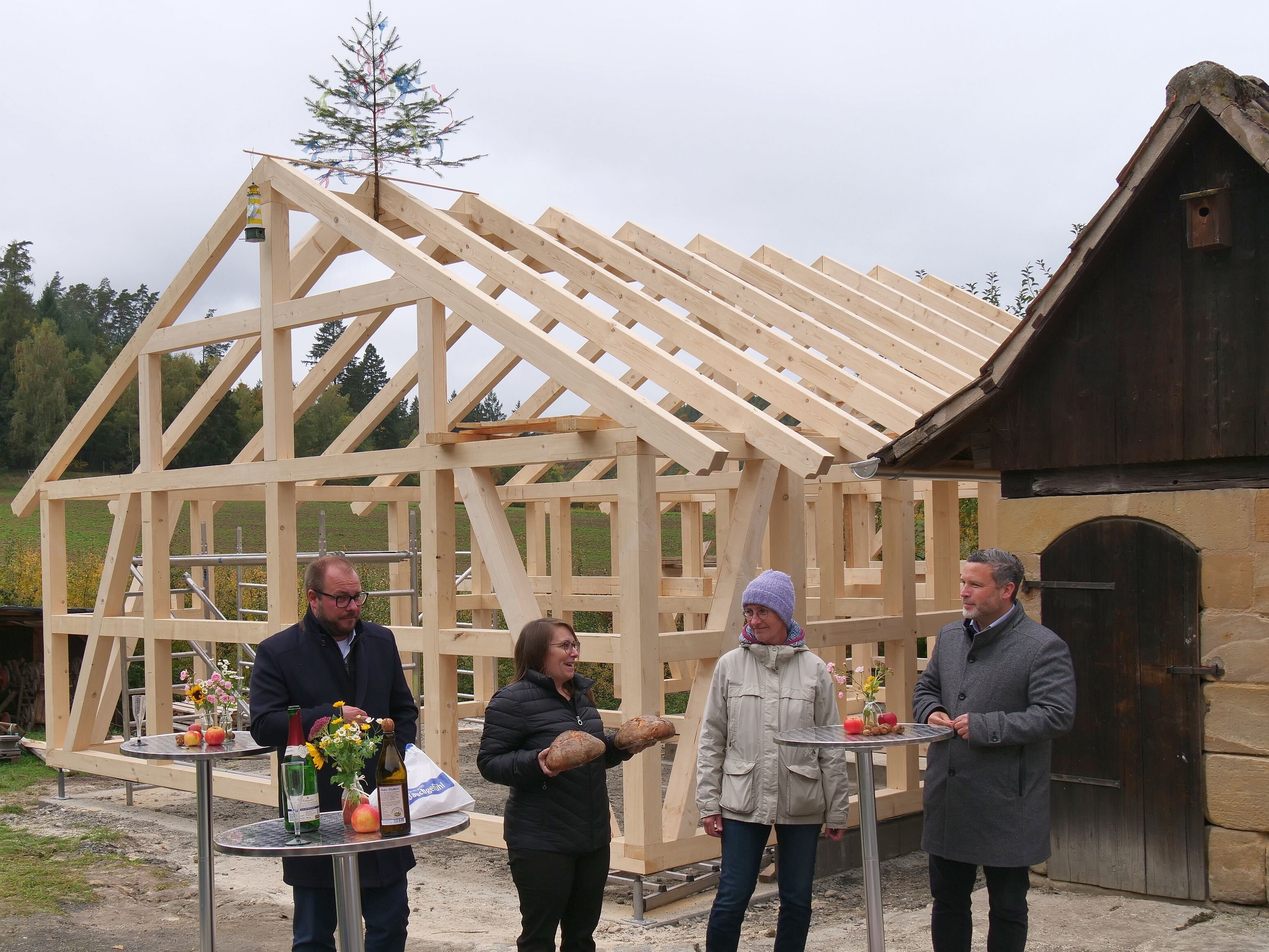 Vier Personen stehen vor dem Holz-Ständer-Gerüst des neuen Gebäudes und unterhalten sich.
