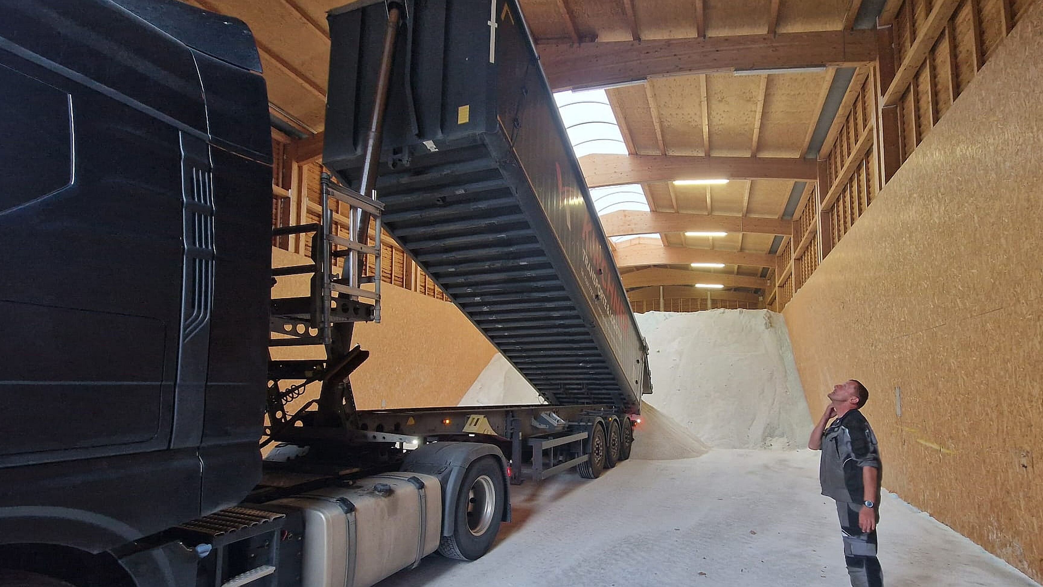 Ein Mann steht neben einem Lastwagen, dessen Ladefläche um 45 Grad nach oben gekippt ist. Hinten vom Lastwagen rieselt Streusalz auf einen großen Berg mit Streusalz.