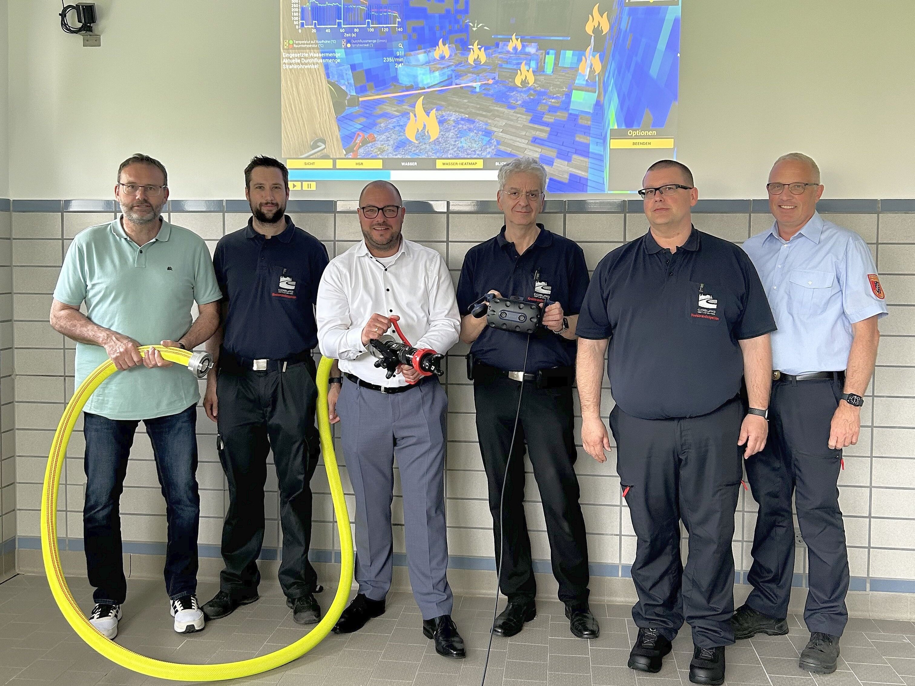 Timo Sommerluksch, Kevin Weinhold, Sebastian Straubel, Stefan Zapf, Michael Hager und Stefan Püls stehen vor einer Wand, auf der im Hintergrund das virtuelle Bild eines brennenden Raumes zu sehen ist.