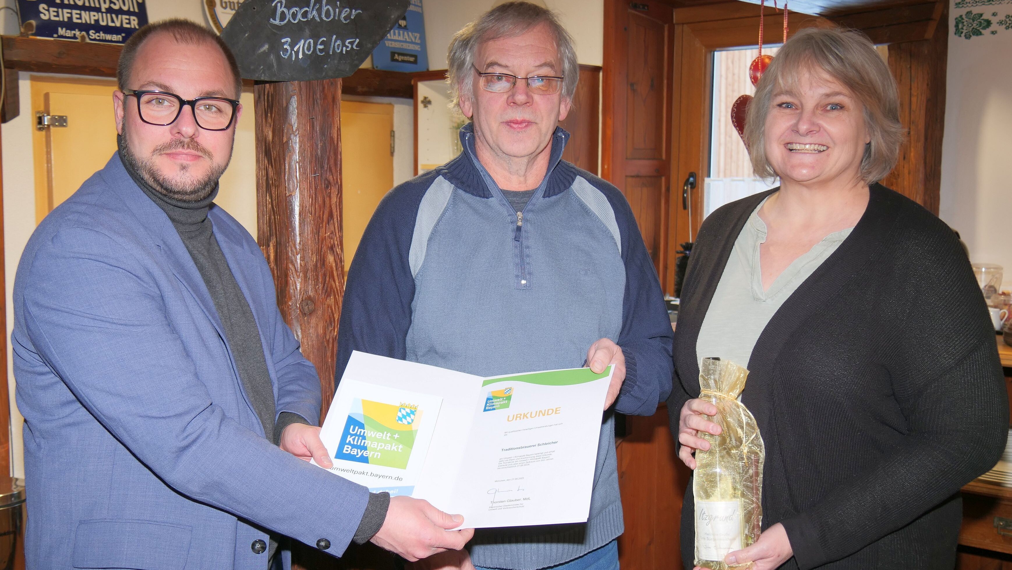 Sebastian Straubel überreicht an Oskar Dölliger eine Urkunde mit dem Logo des bayerischen Umwelt- und Klimapakts. Daneben steht Bürgermeisterin Nina Liebermann mit einer Weinflasche als Präsent in der Hand.