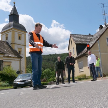 Patrick Mohr steht vor der Kirche in Hassenberg und zeigt auf eine weiß markierte Fläche auf der Straße: Dort befindet sich der Auslösemechanismus für die Ampel.