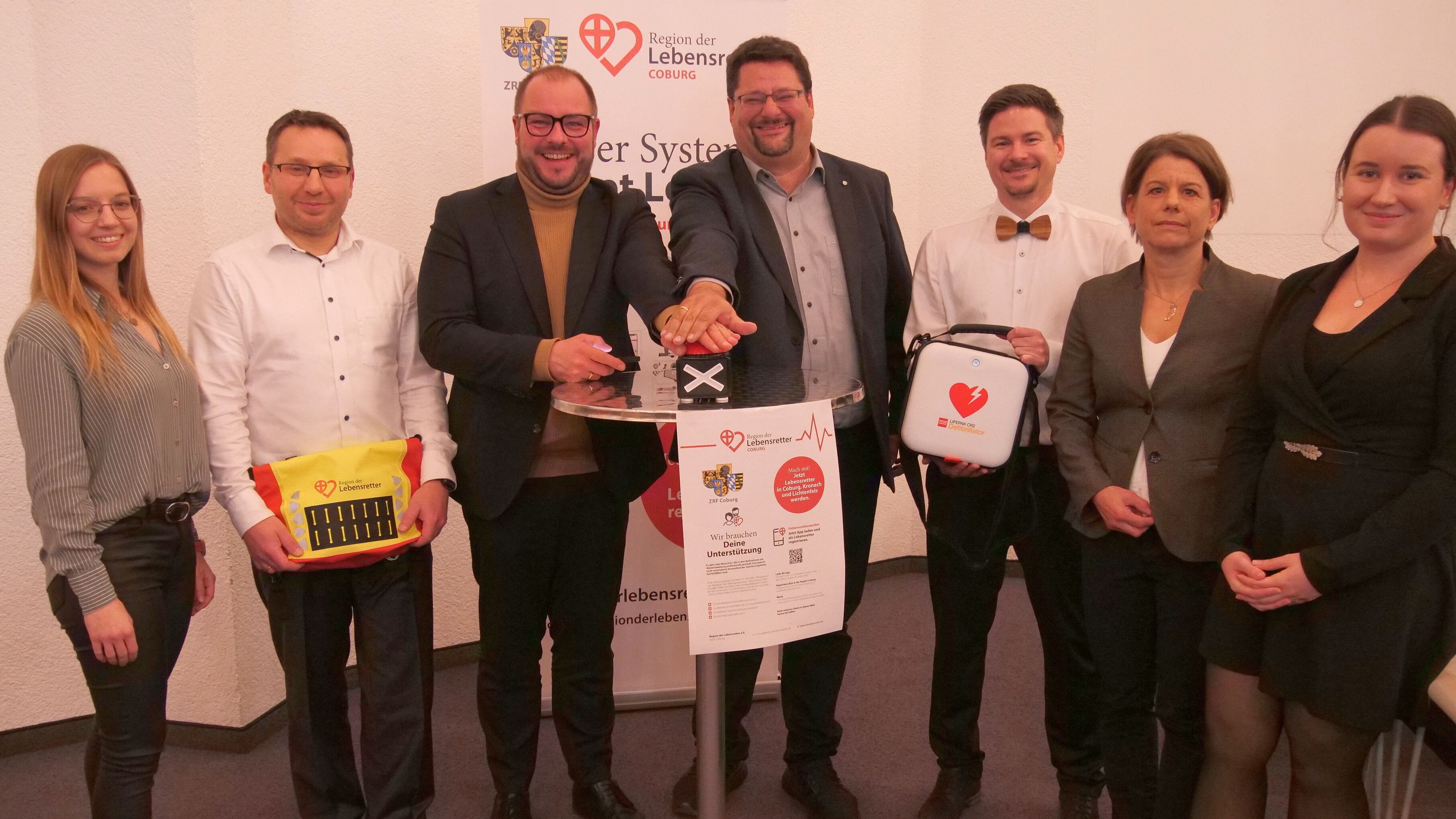 Stefan Wicklein und Sebastian Straubel stehen in der Bildmitte und drücken gemeinsam auf einen Buzzer-Knopf. Daneben stehen verschiedene Personen, die medizische Geräte wie einen Defibrillator und einen Verbandskasten in der Hand halten.
