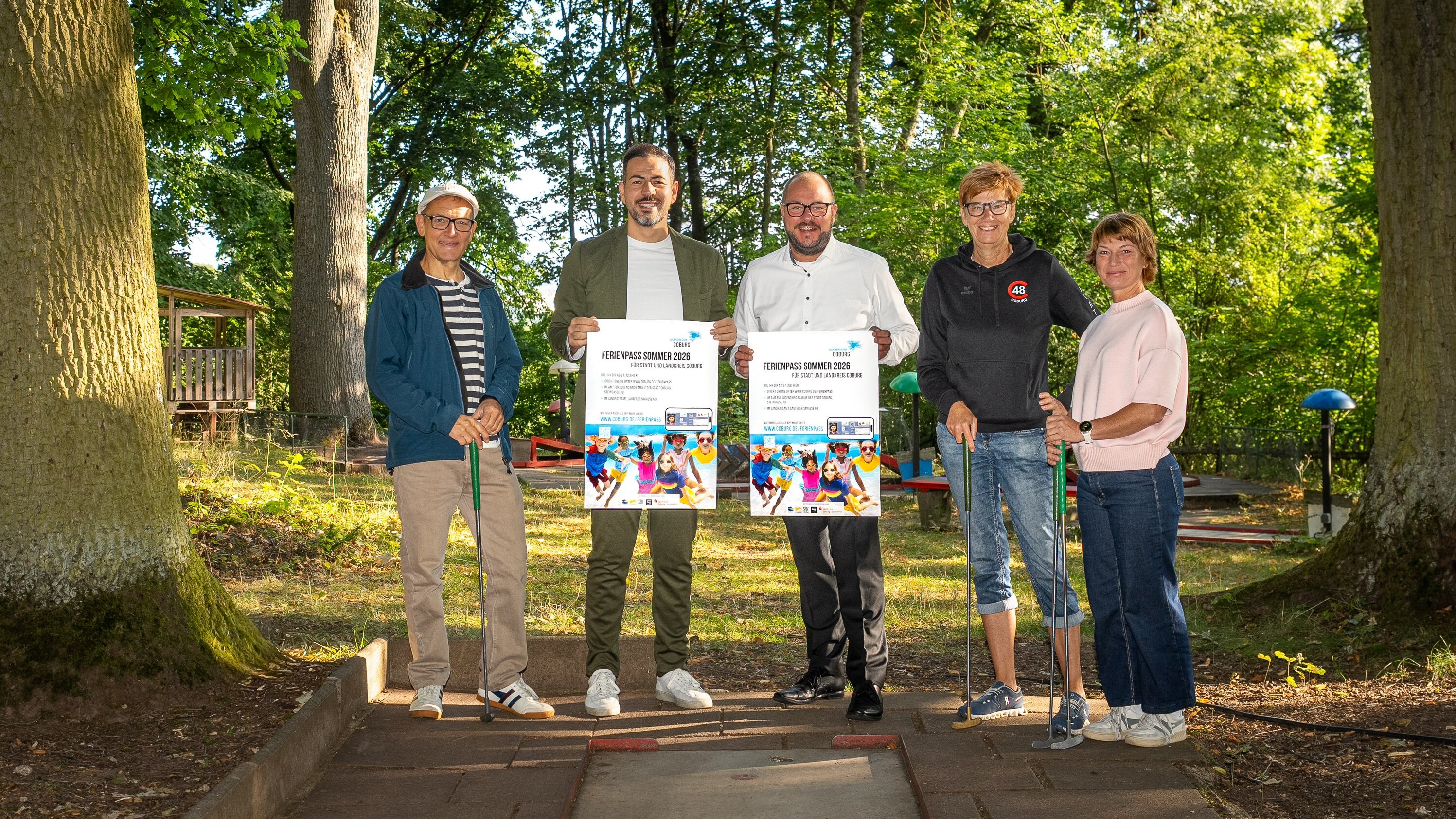 Fünf Personen stehen auf einem Minigolfplatz, zwei von ihnen haben ein Werbeplakat für den Ferienpass in der Hand.