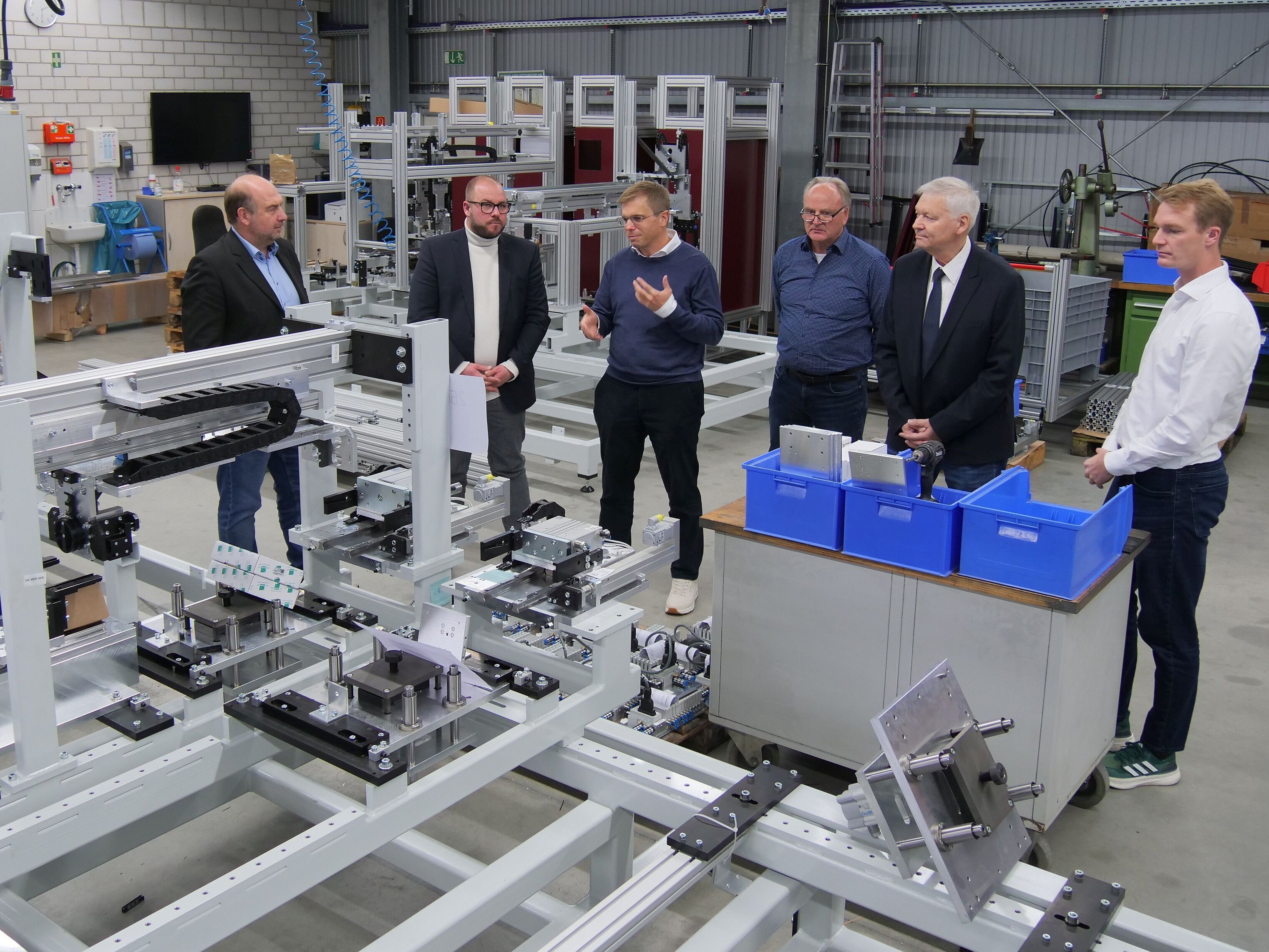 Hinter einer großen Maschine mit unterschiedlichen Aluminiumprofilen, die gerade erst zusammengebaut wird, stehen sechs Personen und unterhalten sich.