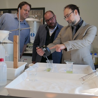 Dr. Sebastian Kamps steckt in einem Labor ein Messgerät in einen Becher mit Flüssigkeit. Sebastian Straubel und Andre Dehler schauen interessiert zu.