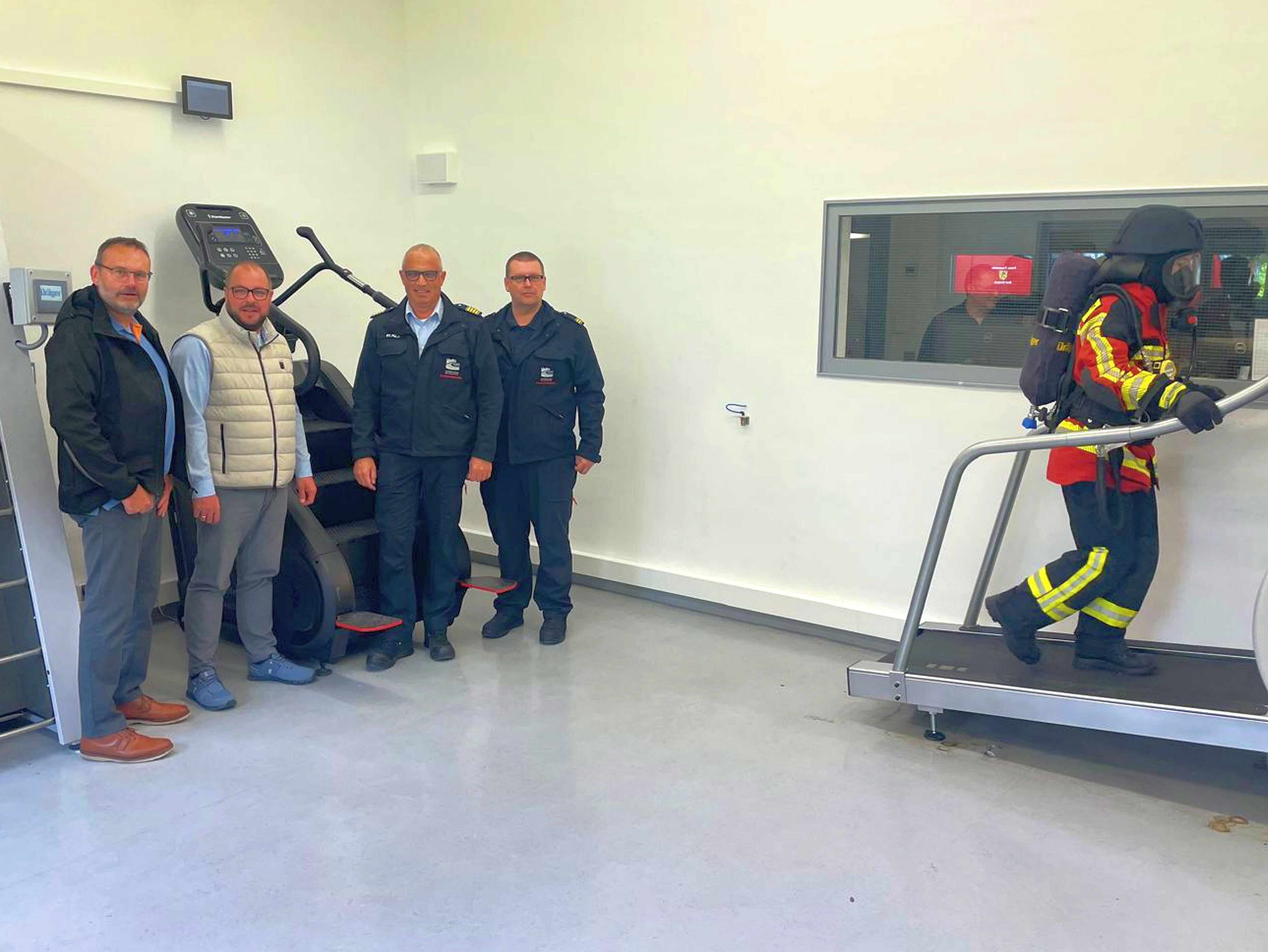 Ein Feuerwehrmann mit Atemschutzausrüstung trainiert auf einem Laufband. Mehrere Personen in Zivil- und Feuerwehr-Kleidung schauen ihm zu.