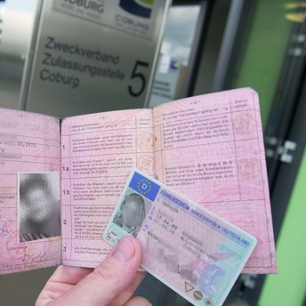 Eine Hand hält einen alten rosafarbenen sowie einen neuen Kartenführerschein. Im Hintergrund ist der Eingang zur Zulassungsstelle Coburg zu sehen.
