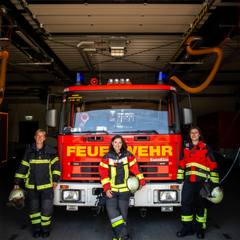Drei Feuerwehrfrauen stehen in Uniform vor einem Feuerwehrfahrzeug 
