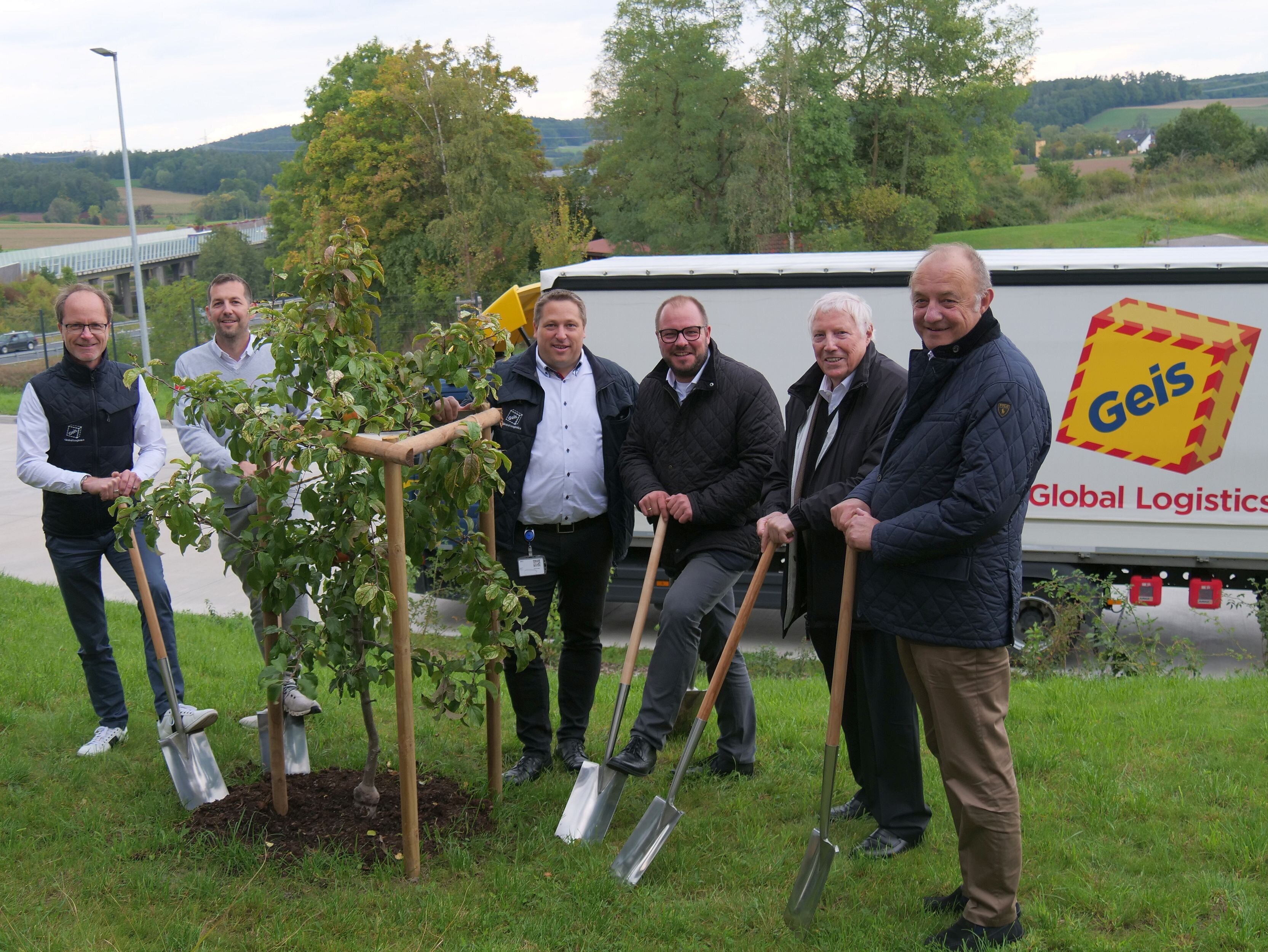 Sechs Personen stehen mit jeweils einem Spaten in der Hand vor einem frisch gepflanzten Baum. Im Hintergrund ist ein Lkw mit dem Schriftzug der Firma Geis zu sehen.