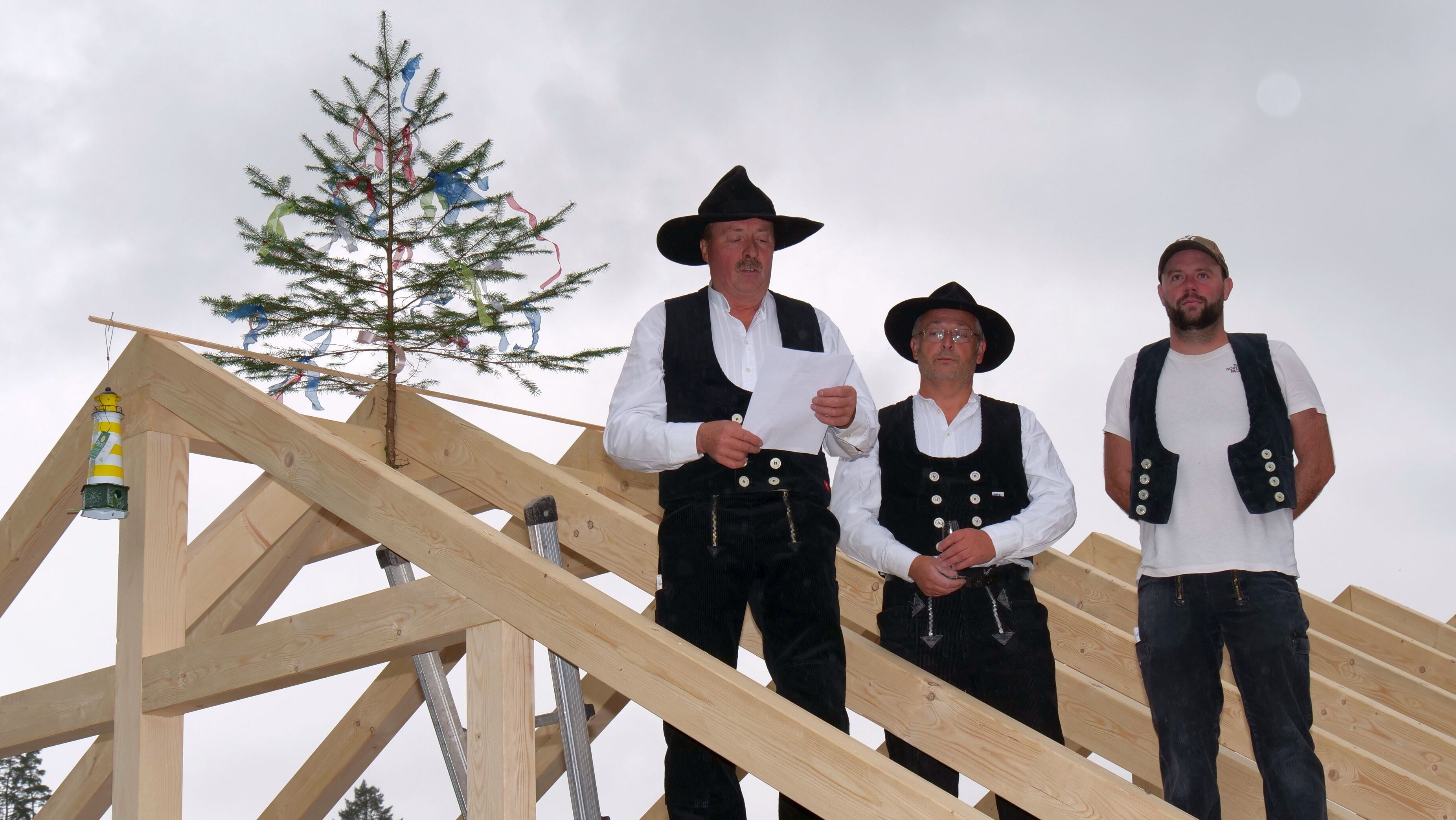 Peter Kraft steht mit zwei Zimmermanns-Kollegen in typischer Zimmermannskleidung auf der Dachkonstruktion und hält den Richtspruch. Im Hinter steht auf der Dach-Spitze eine kleine Fichte.