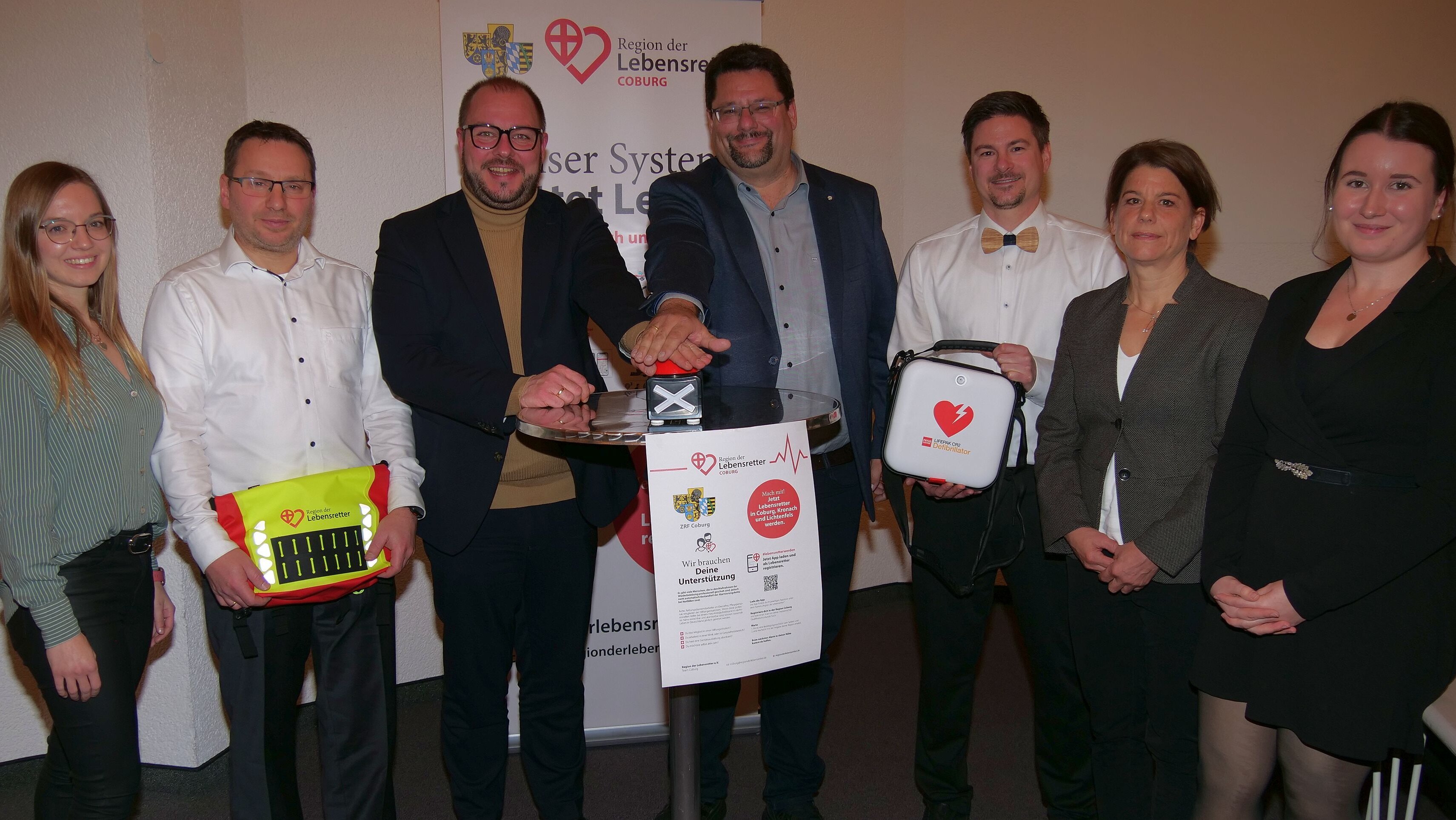 Stefan Wicklein und Sebastian Straubel stehen in der Bildmitte und drücken gemeinsam auf einen Buzzer-Knopf. Daneben stehen verschiedene Personen, die medizische Geräte wie einen Defibrillator und einen Verbandskasten in der Hand halten.