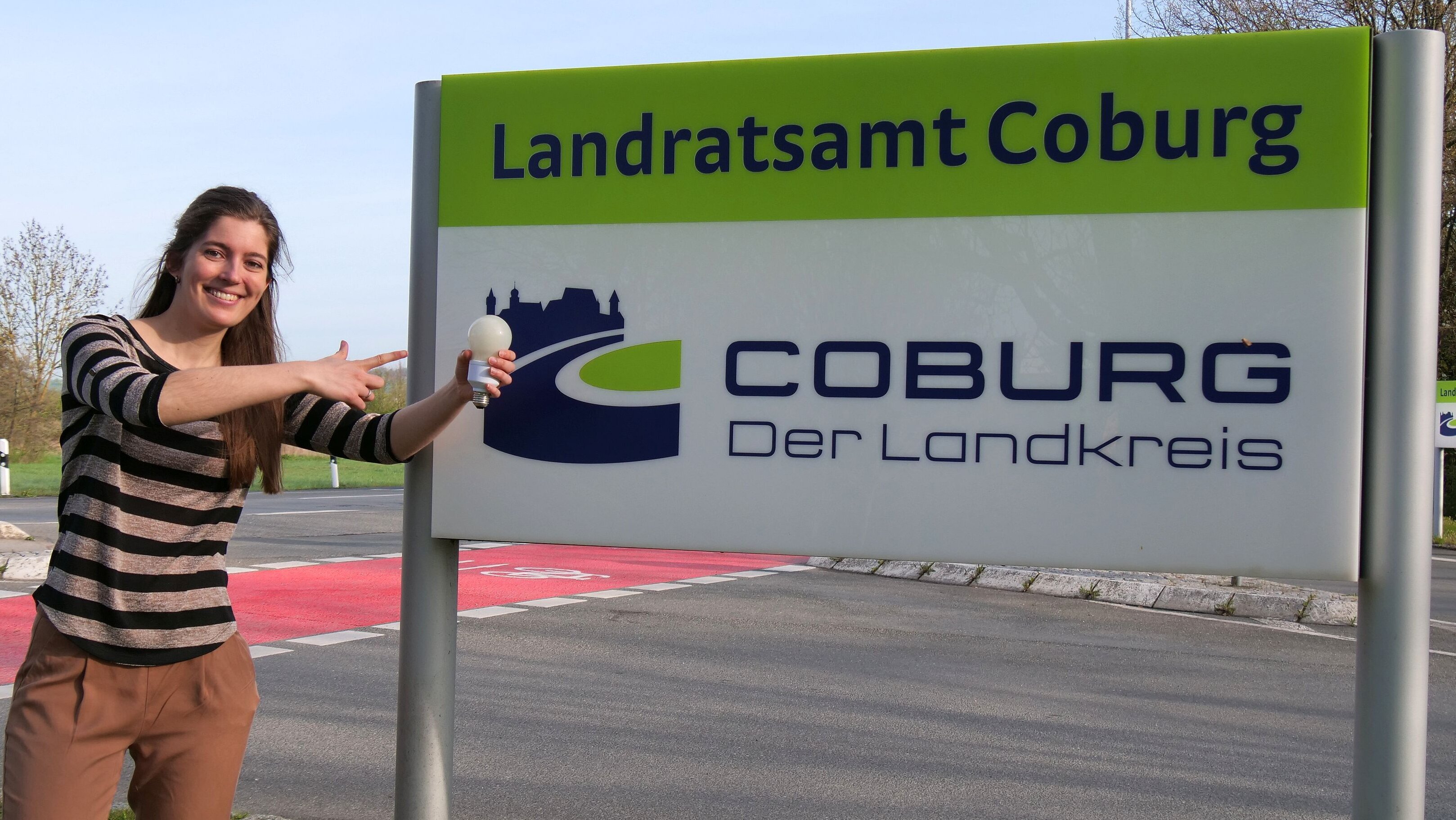 Klimaschutzmanagerin Franziska Dennstädt steht vor einem Schild mit der Aufschrift "Landratsamt Coburg". Sie zeigt mir der rechten Hand auf eine Energiesparbirne, die sie in der rechten Hand hält.