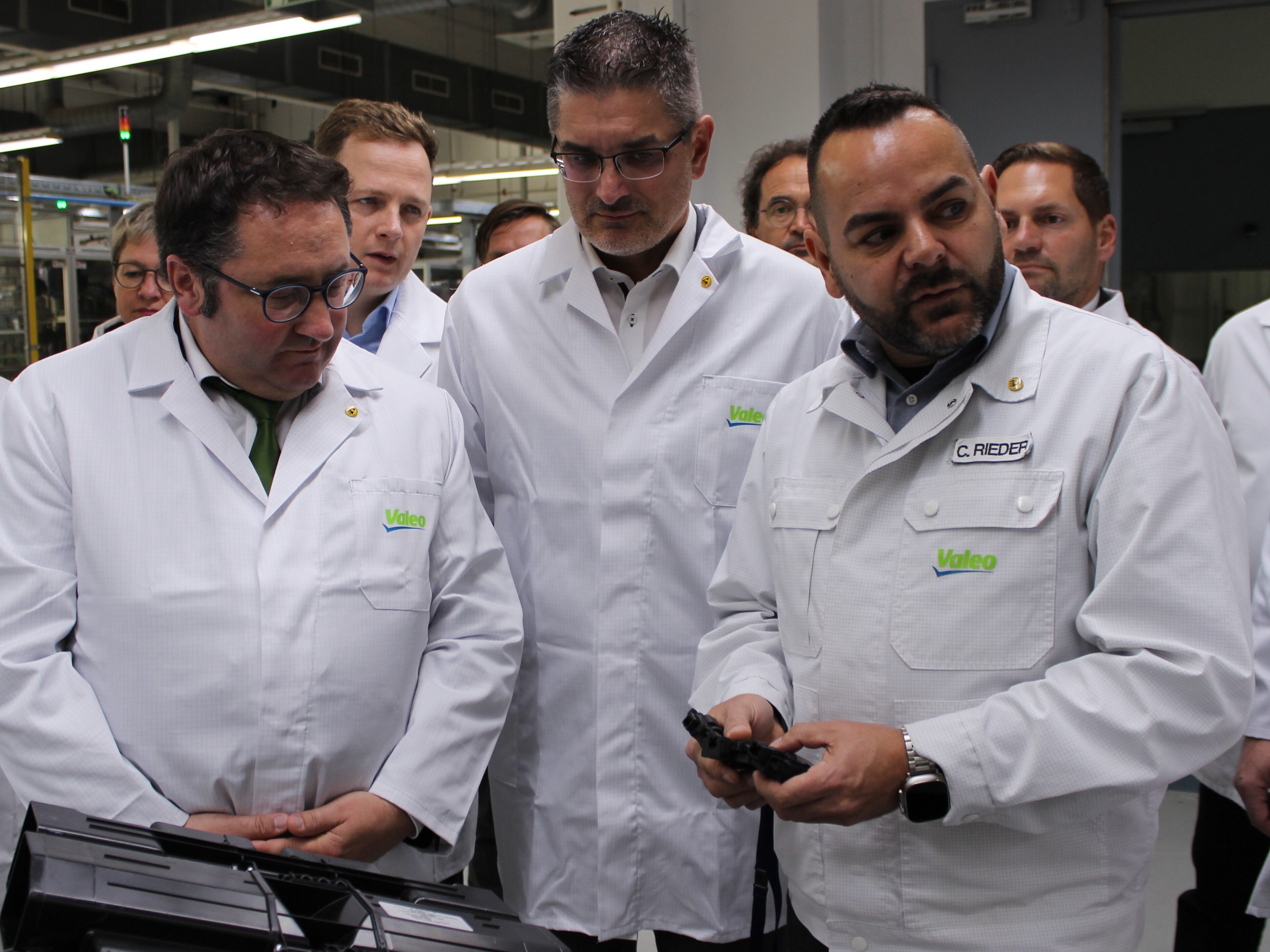 Bekleidet mit weißen Schutzjacken begutachten Staatssektretär Tobias Gotthardt und weitere Personen schwarze Kunststoffteile, die bei der Herstellung von Fahrzeugkomponenten benötigt werden.