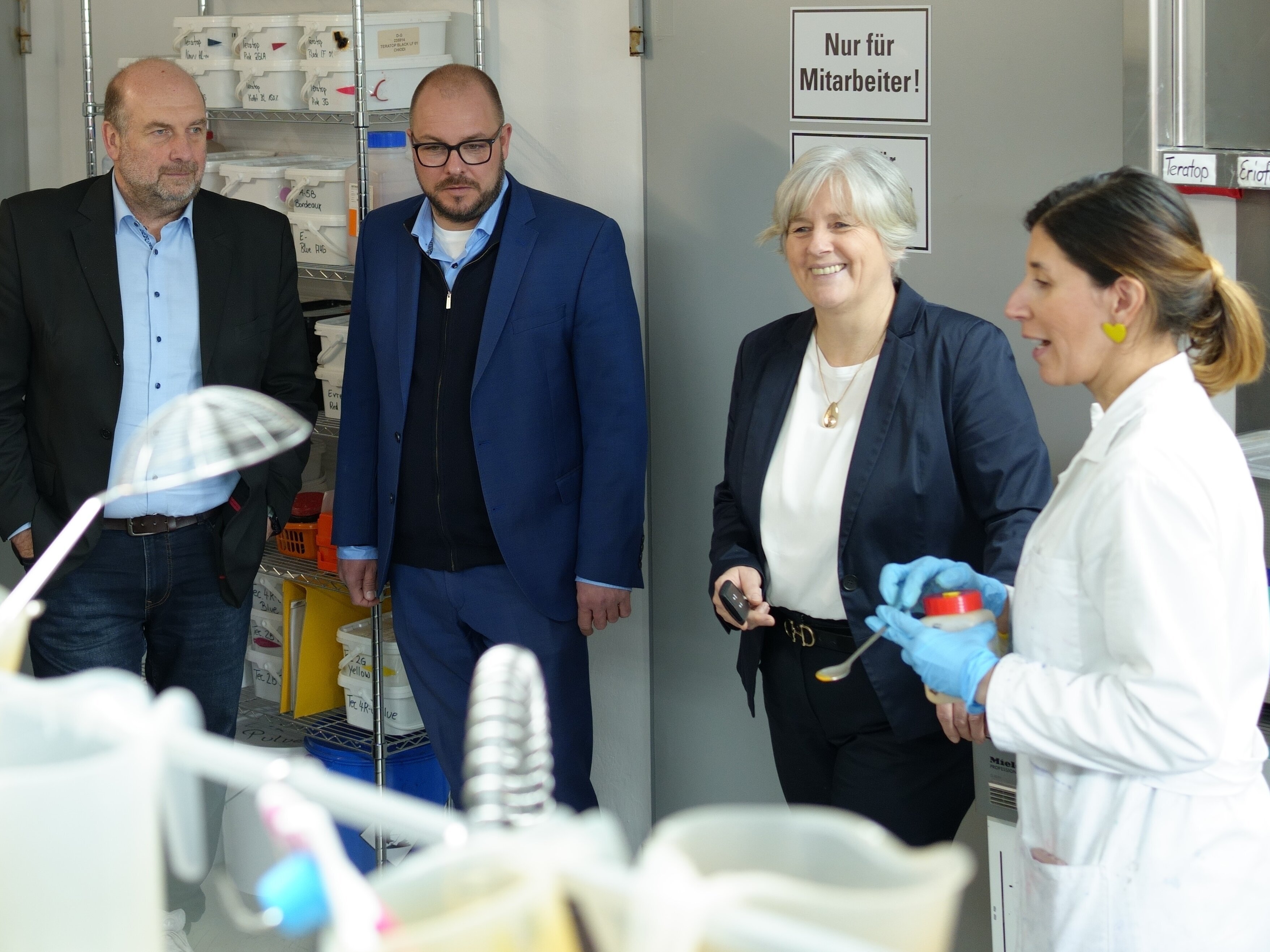 Vier Personen stehen in einem Labor. Rechts eine Frau in einem weißen Laborkittel mit einer Chemikaliendose in der Hand. Sie erklärt, was sie gerade macht.