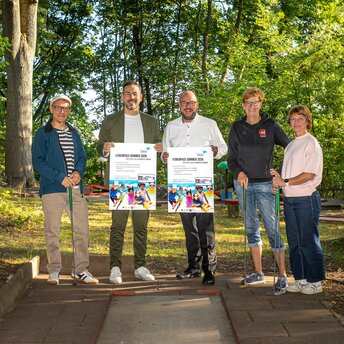 Vier Personen stehen auf einem Minigolfplatz, zwei davon haben ein Werbeplakat für den Ferienpass in der Hand.